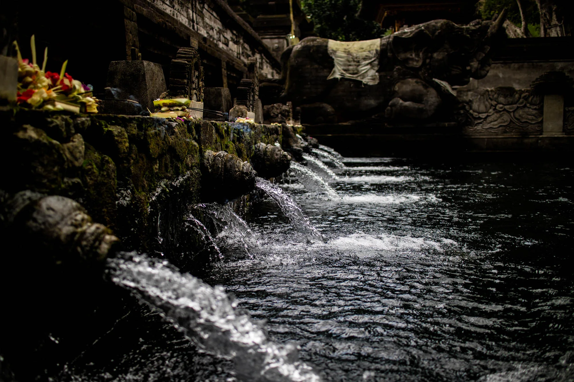 インドネシアのバリ島で撮影された写真です。暗く、神秘的な雰囲気が漂っています。手前には、苔むした石造りの壁があり、そこからいくつもの水が勢いよく流れ出ています。水の流れは滝のようにも見え、水面は光を反射してきらきらと輝いています。壁の上には、花や供え物が置かれており、神聖な場所であることを示しています。背景には、石像の一部が見え、バリの寺院や聖域の一部であることが伺えます。全体的に、バリの伝統と精神性が感じられる、静かで神聖な場所を捉えた写真です。
