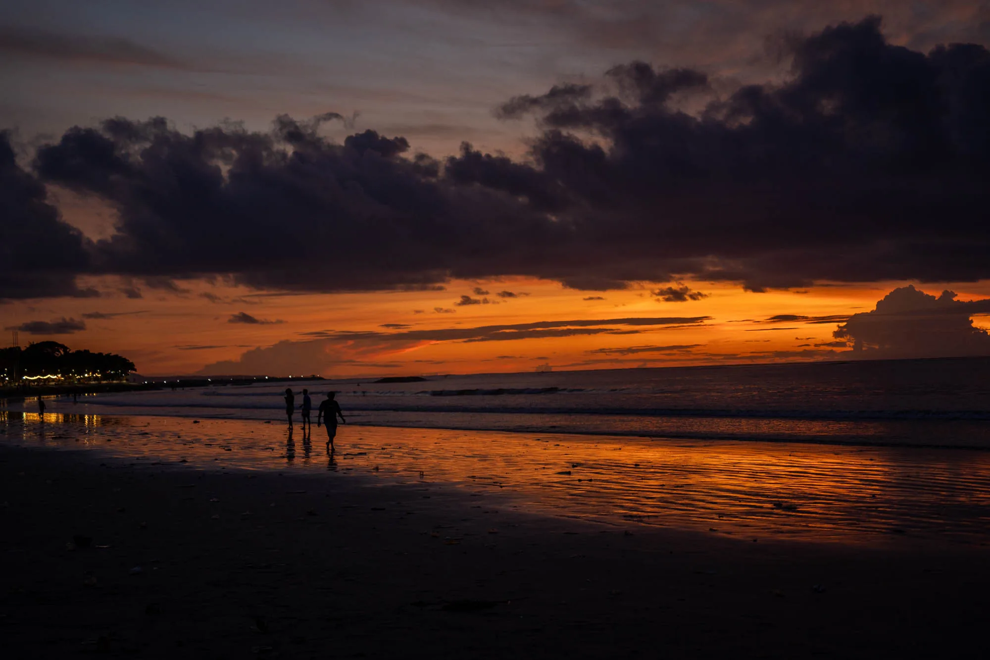 インドネシアのバリで撮影されたと思われる写真です。空は夕焼けで、オレンジ色と紫色のグラデーションが美しいです。厚い雲が空を覆い、ドラマチックな雰囲気を醸し出しています。海岸線には穏やかな波が打ち寄せ、夕焼けの色を反射して輝いています。砂浜には人影がいくつか見え、シルエットになっています。彼らは海辺を散歩しているようです。背景には街灯が灯る建物や木々が見え、海岸沿いの風景であることを示唆しています。全体的に、夕暮れ時の穏やかで美しい光景が捉えられています。
