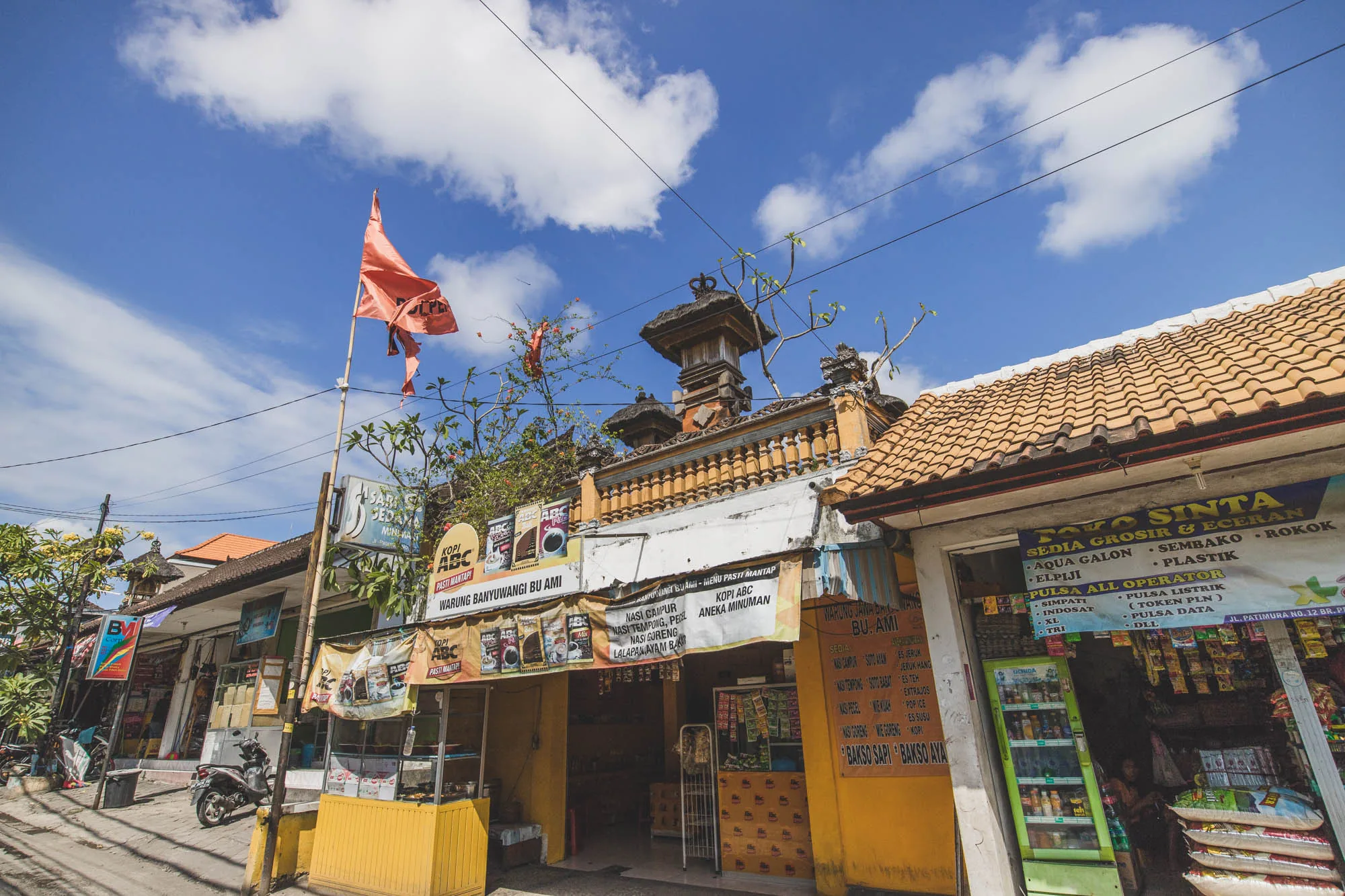 インドネシアのバリ島で撮影された写真です。青空に白い雲が浮かび、その下にはオレンジ色の旗がはためいています。通り沿いには、鮮やかな色彩の商店が軒を連ねています。左手には Warung Banyuwangi Bu Ami という名前の飲食店があり、メニューが看板に書かれています。その隣には、Toko Sinta という商店があり、食料品や日用品を扱っているようです。店の前にはスクーターが停められています。全体的に活気があり、ローカルな雰囲気が感じられる写真です。