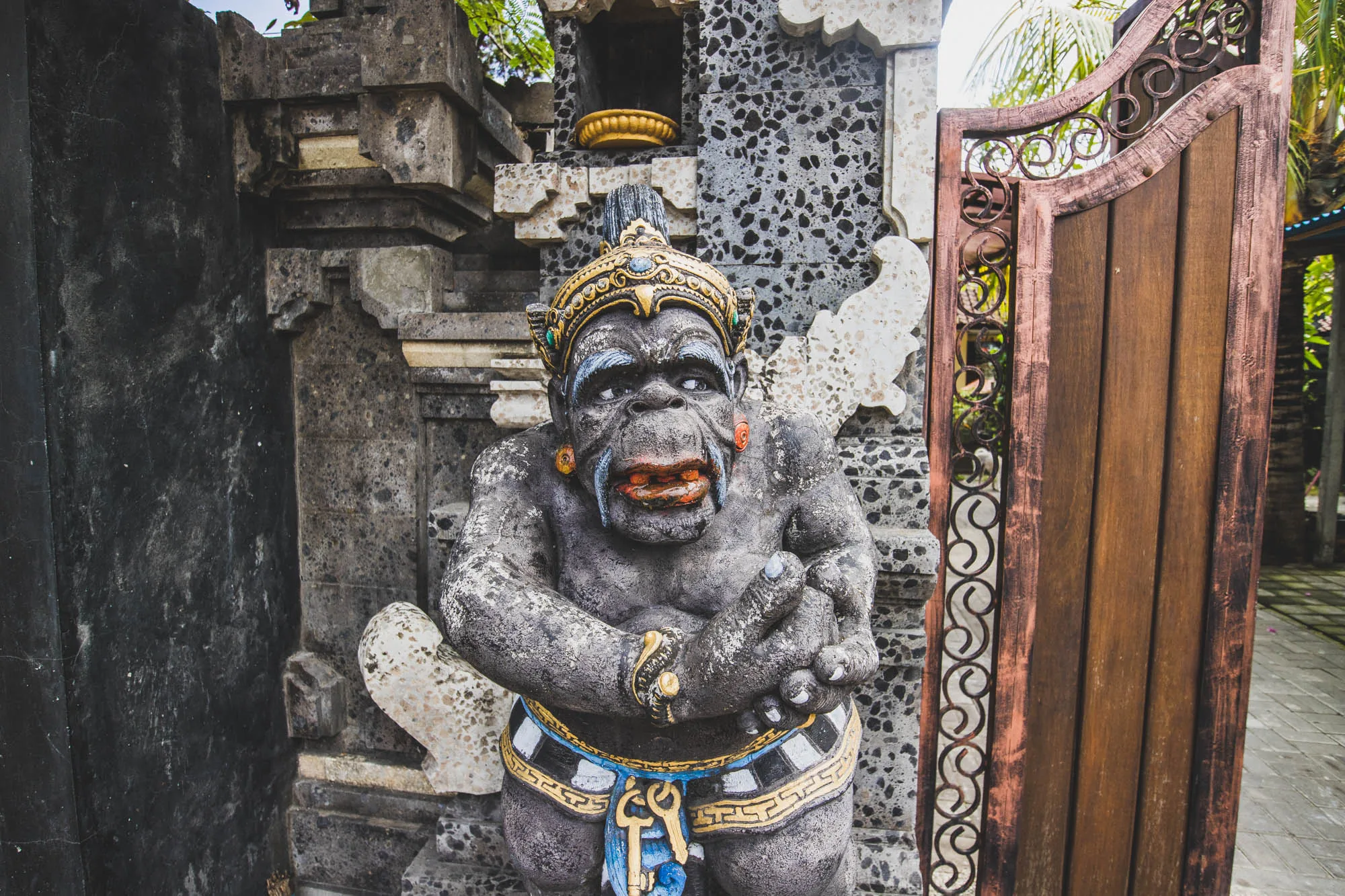 写真はインドネシアのバリ島で撮影されたもので、石造りの像と門が写っています。像は、猿のような顔立ちで、王冠をかぶり、腰には装飾が施されています。両手を握りしめ、やや前かがみになっています。像の背景には、複雑な模様が施された石垣があり、その上には小さな祠のようなものが置かれています。像の右側には木製の門があり、鉄製の装飾が施されています。門の向こうには緑豊かな植物が見え、周囲の熱帯の雰囲気を醸し出しています。像と門はやや古びた印象で、バリの伝統的な建築様式と文化を感じさせます。全体的に、神秘的でエキゾチックな雰囲気の写真です。