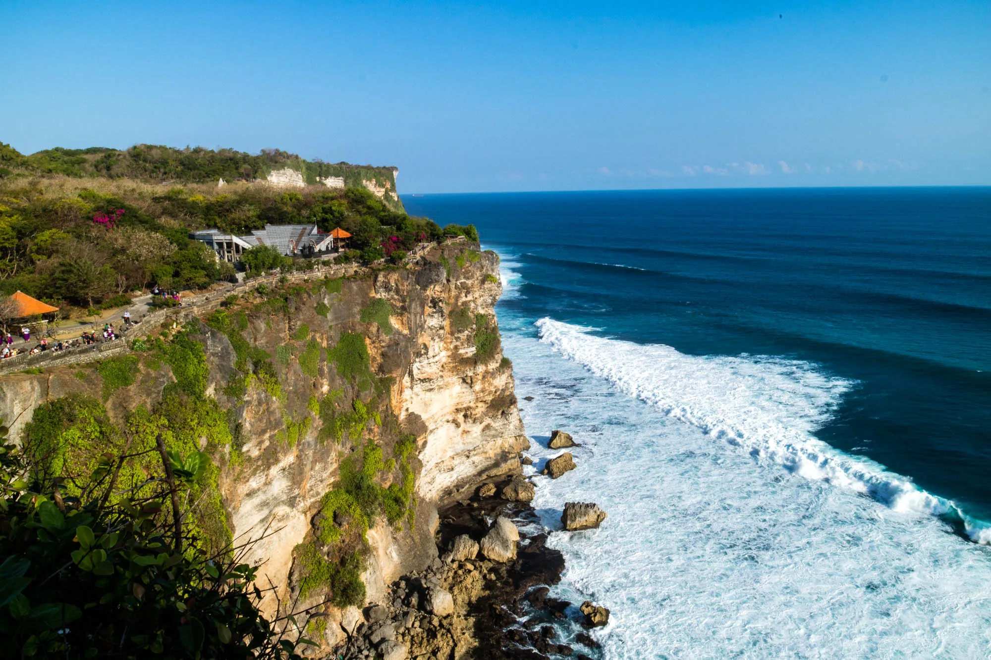 インドネシアのバリ島で撮影された写真です。写真には、切り立った崖と、その下に広がる青い海が写っています。崖の上には緑豊かな植物が生い茂り、いくつかの建物が見えます。崖沿いには遊歩道のようなものがあり、人が歩いている様子が確認できます。空は晴れ渡り、太陽光が海面に反射して輝いています。波は白く砕け、海岸線に沿って打ち寄せています。全体的に、壮大で美しい自然の風景が捉えられています。崖の質感や植物の細部、海の色のグラデーションなど、細部に至るまで鮮明に描写されており、臨場感があります。静かで穏やかな雰囲気が感じられます。