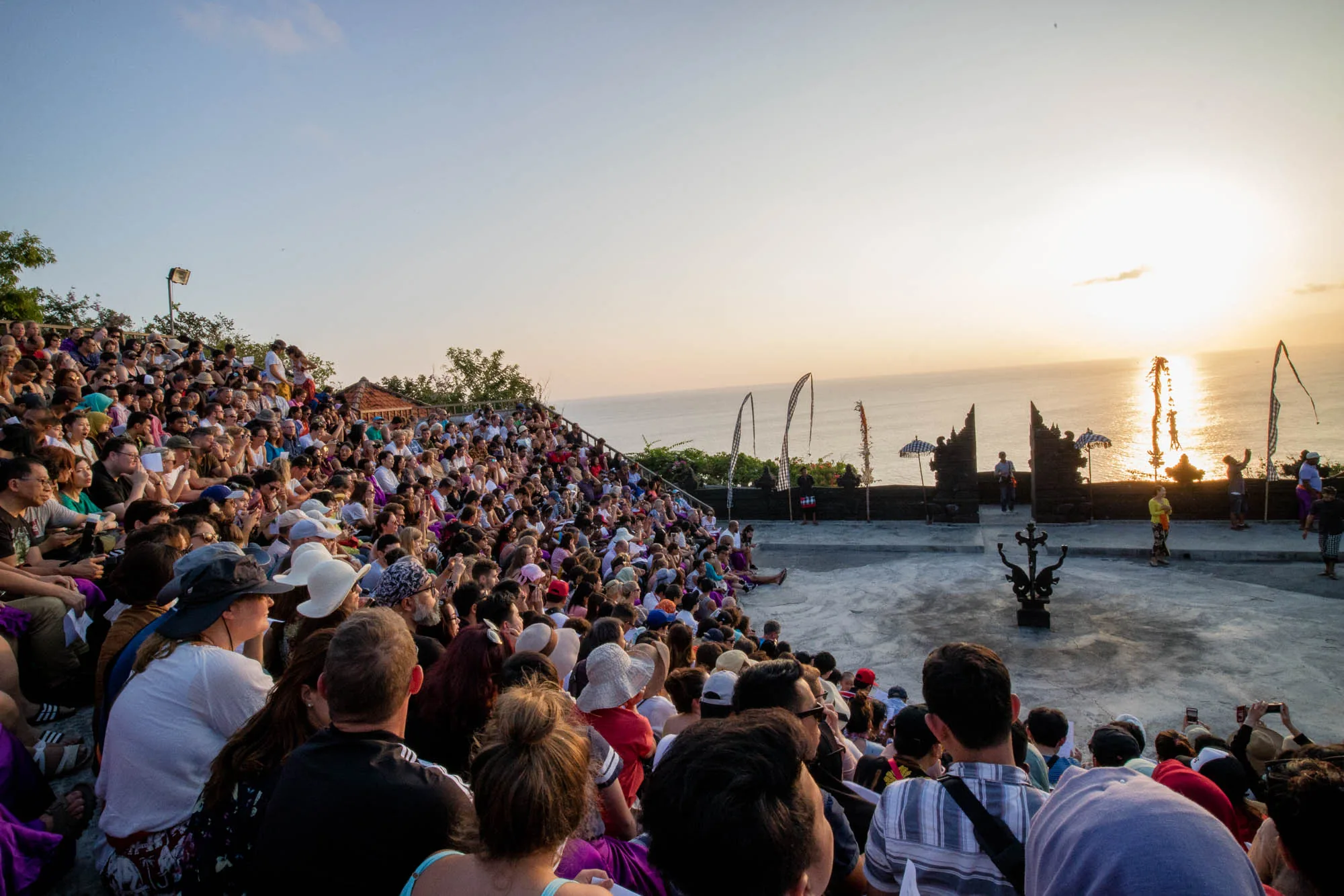 インドネシアのバリで撮影されたと思われる写真です。夕暮れ時の海を背景に、階段状の観客席に多くの人々が集まっています。観客はさまざまな服装をしており、帽子をかぶっている人も見られます。彼らの視線の先には、舞台のような場所があり、装飾的な門や旗のようなものが立っています。舞台には数人の人が立っており、何らかのパフォーマンスが行われるのか、あるいは終わったのかは不明です。夕日の光が海面に反射してきらめき、空はオレンジ色に染まっています。観客の中にはスマートフォンで写真を撮っている人もおり、この美しい光景を記録しようとしている様子が伺えます。全体的に、バリの夕日を背景にしたイベントの様子を捉えた、活気のある写真です。