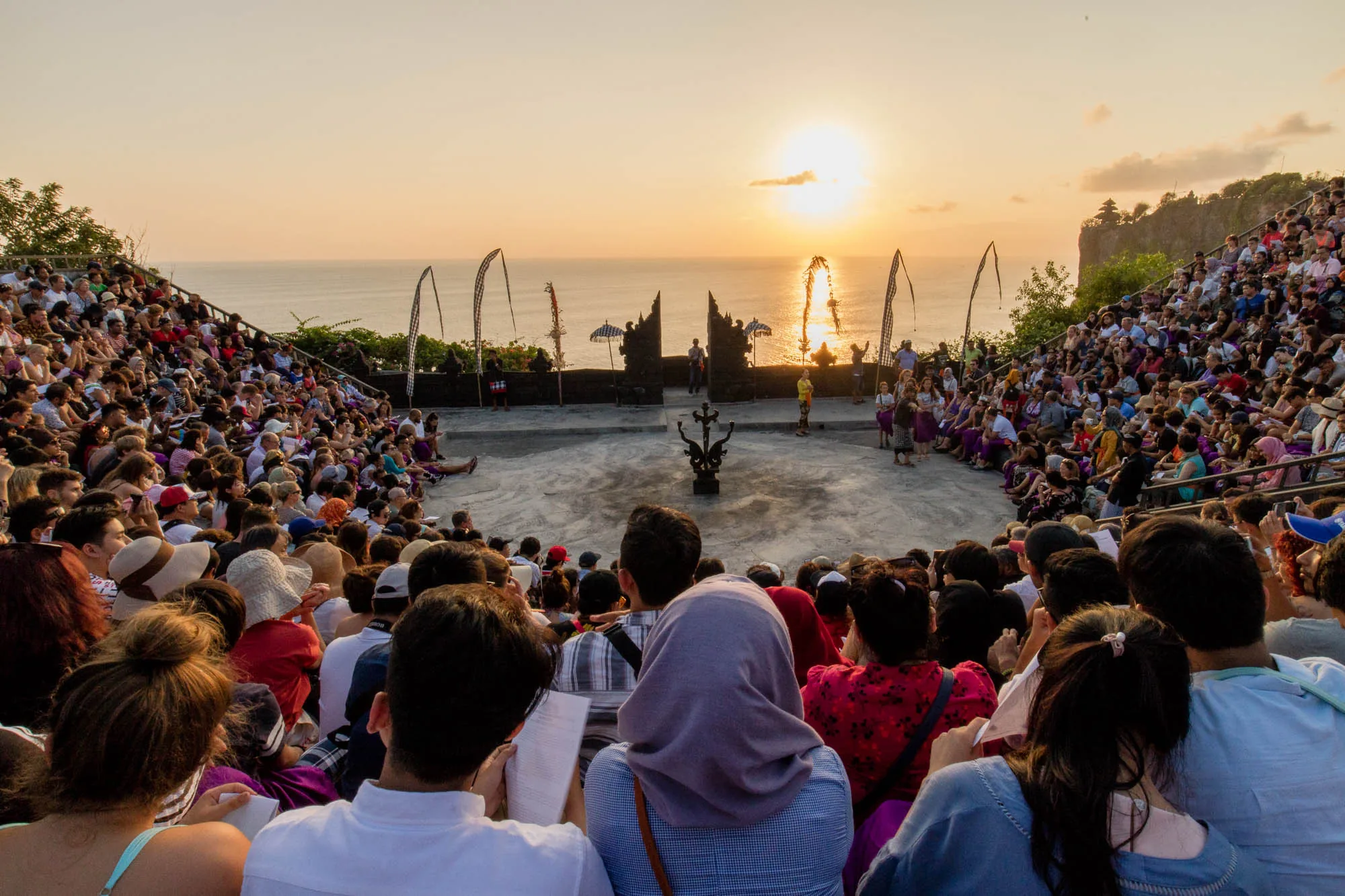 この写真はインドネシアのバリ島で撮影されたもので、夕暮れ時の雰囲気が伝わってくる。階段状の客席には大勢の観客が座っており、年齢層も服装も様々で、観光客が多い様子が見て取れる。舞台の中央には装飾的なオブジェが置かれ、その奥には海と夕日が見える。伝統的な門構えや旗が建てられており、バリの寺院の雰囲気を感じさせる。舞台上には、民族衣装を着た人々が数名おり、これから伝統芸能が始まることを予感させる。全体的に、夕日を背景にしたバリの伝統芸能を楽しむ人々の様子が写し出されており、穏やかで美しい風景が印象的である。