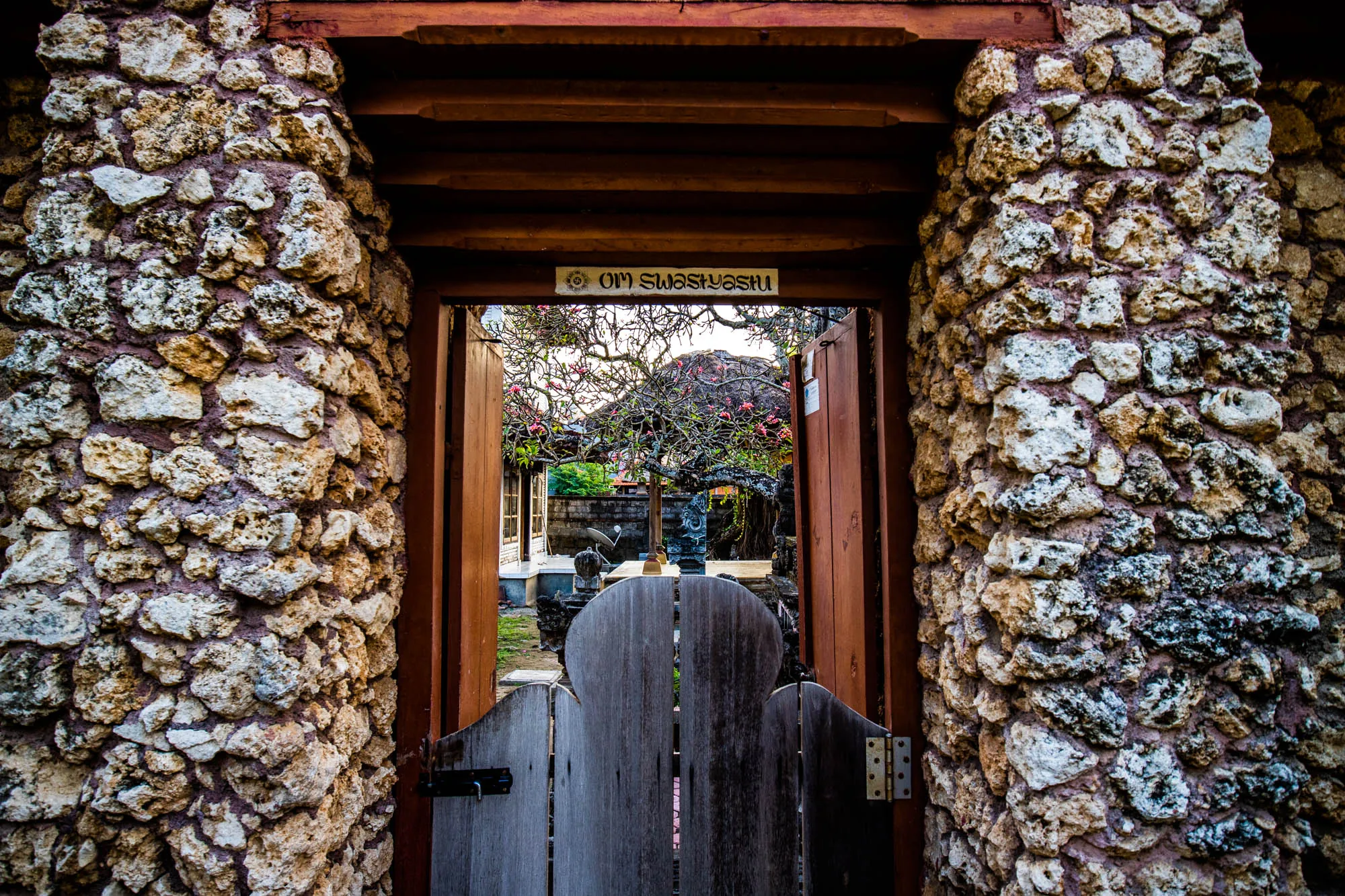 インドネシアのバリで撮影された写真です。写真は、石造りの門から庭を覗いた様子を捉えています。門の柱は大小さまざまな岩を積み重ねて作られており、自然な風合いが感じられます。門扉は木製で、中央部分が特徴的な形状に切り取られています。門の上部には木製の梁があり、「OM SWASTYASTU」と書かれたプレートが掲げられています。「OM SWASTYASTU」はサンスクリット語で「神のご加護がありますように」という意味です。門の向こうには、緑豊かな庭が広がっており、木々や石像が見えます。背景には建物の一部が見え、バリの伝統的な建築様式を垣間見ることができます。全体的に落ち着いた色調で、静かで平和な雰囲気が漂っています。