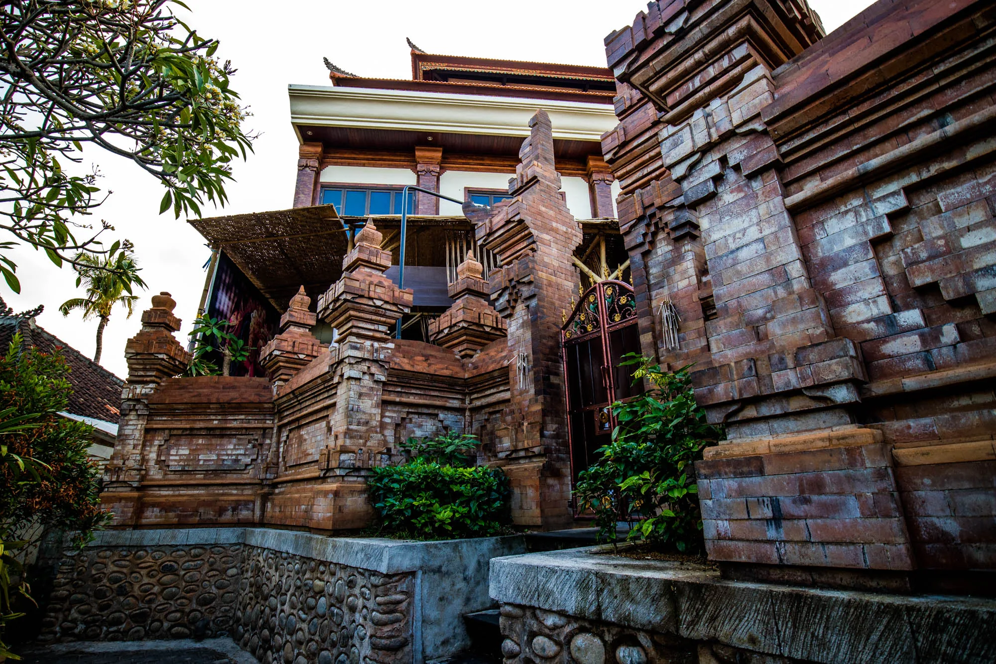 インドネシアのバリ島で撮影された写真です。写っているのは、レンガ造りの伝統的な建築物です。建物は、バリの独特なスタイルで装飾されており、細部まで丁寧に作り込まれています。レンガの色合いは、赤茶色を基調としており、歴史を感じさせる風合いです。建物の周囲には緑豊かな植物が植えられており、自然と建築が調和した美しい景観を作り出しています。石垣の塀と植物によって、建物の重厚さが際立っています。奥には屋根の形が特徴的な建物が見え、バリらしい雰囲気を醸し出しています。