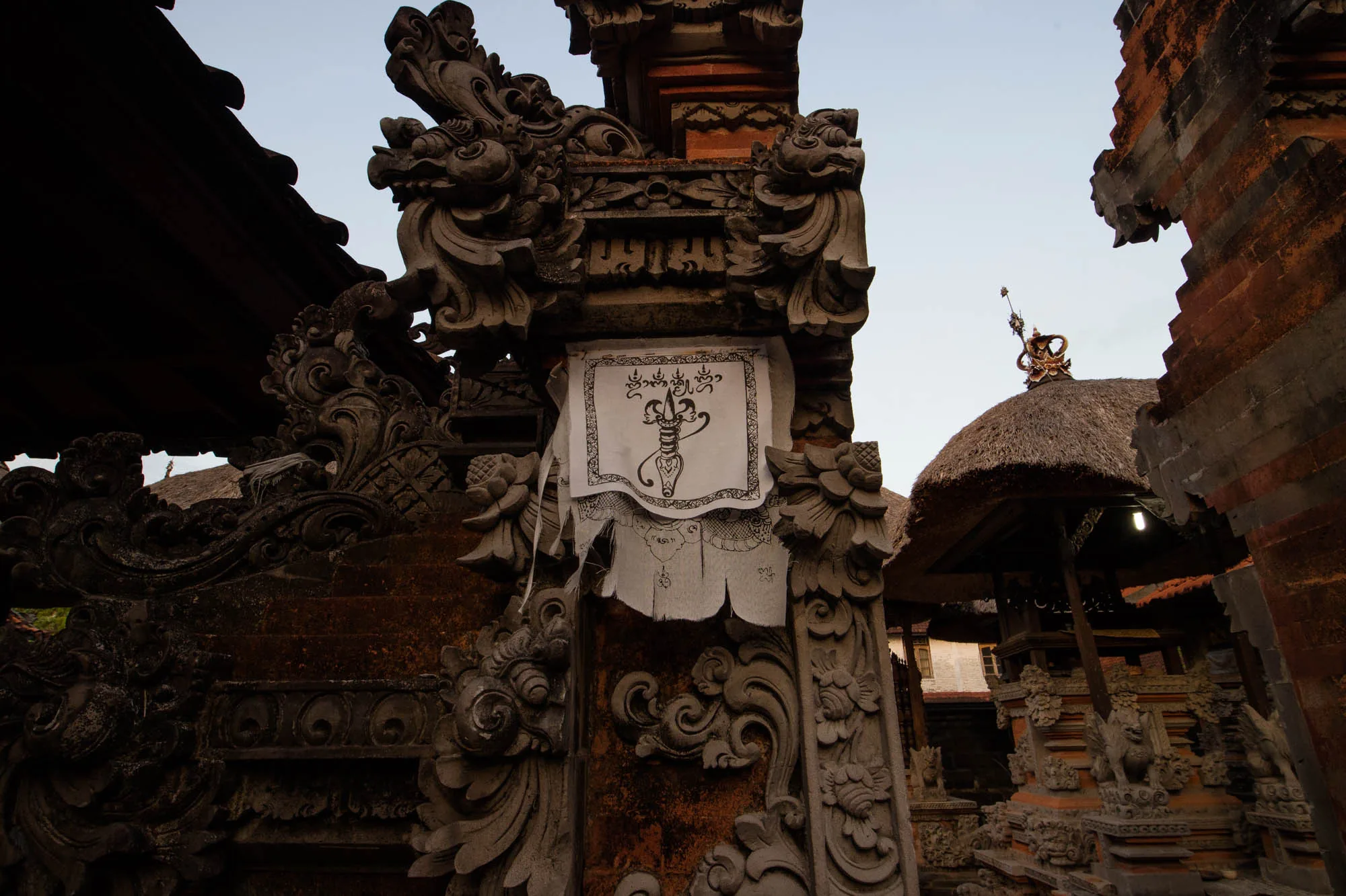 この写真はインドネシアのバリ島で撮影されたもので、寺院の一部を写しています。石造りの門柱には細かく彫刻が施されており、神話的な生き物や植物を思わせる装飾が見られます。門柱に掛けられた白い布には、黒い線で描かれた模様が施されています。これは宗教的な意味を持つ図案かもしれません。  背景には茅葺屋根の建物が見え、これもまた寺院の一部であると考えられます。全体的に落ち着いた色調で、神聖な場所の静謐な雰囲気が伝わってきます。石の質感や彫刻のディテールが、バリの伝統的な建築様式を際立たせています。