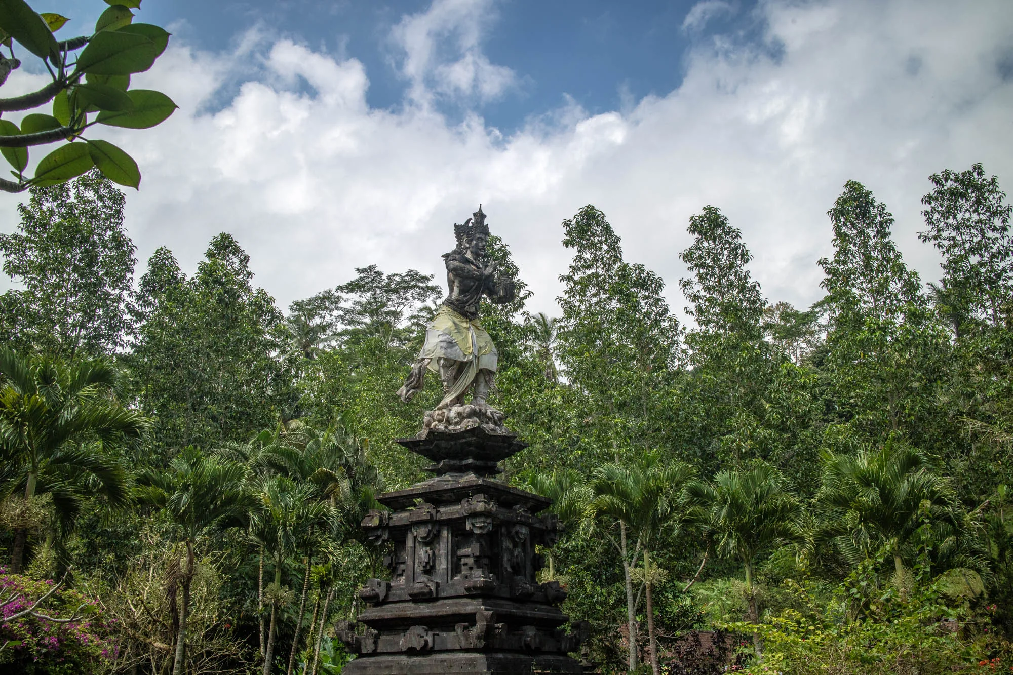 インドネシアのバリで撮影された写真です。画面の中央には、精巧な彫刻が施された石像がそびえ立っています。石像は伝統的な衣装を身につけ、特徴的な冠をかぶっており、神聖な雰囲気を醸し出しています。石像は段になった台座の上に設置されており、その台座にも複雑な模様が刻まれています。  背景には、鬱蒼とした緑が生い茂っています。様々な種類の木々や植物が密集し、熱帯のジャングルのような雰囲気を作り出しています。空は青く、白い雲が浮かんでおり、全体的に明るく開放的な印象を与えます。  写真全体から、バリの豊かな文化と自然が感じられます。石像の荘厳さと、周囲の自然の美しさが調和し、神秘的で魅力的な風景を作り出しています。落ち着いた色調と静寂な雰囲気は、見る人をバリの精神世界へと誘います。