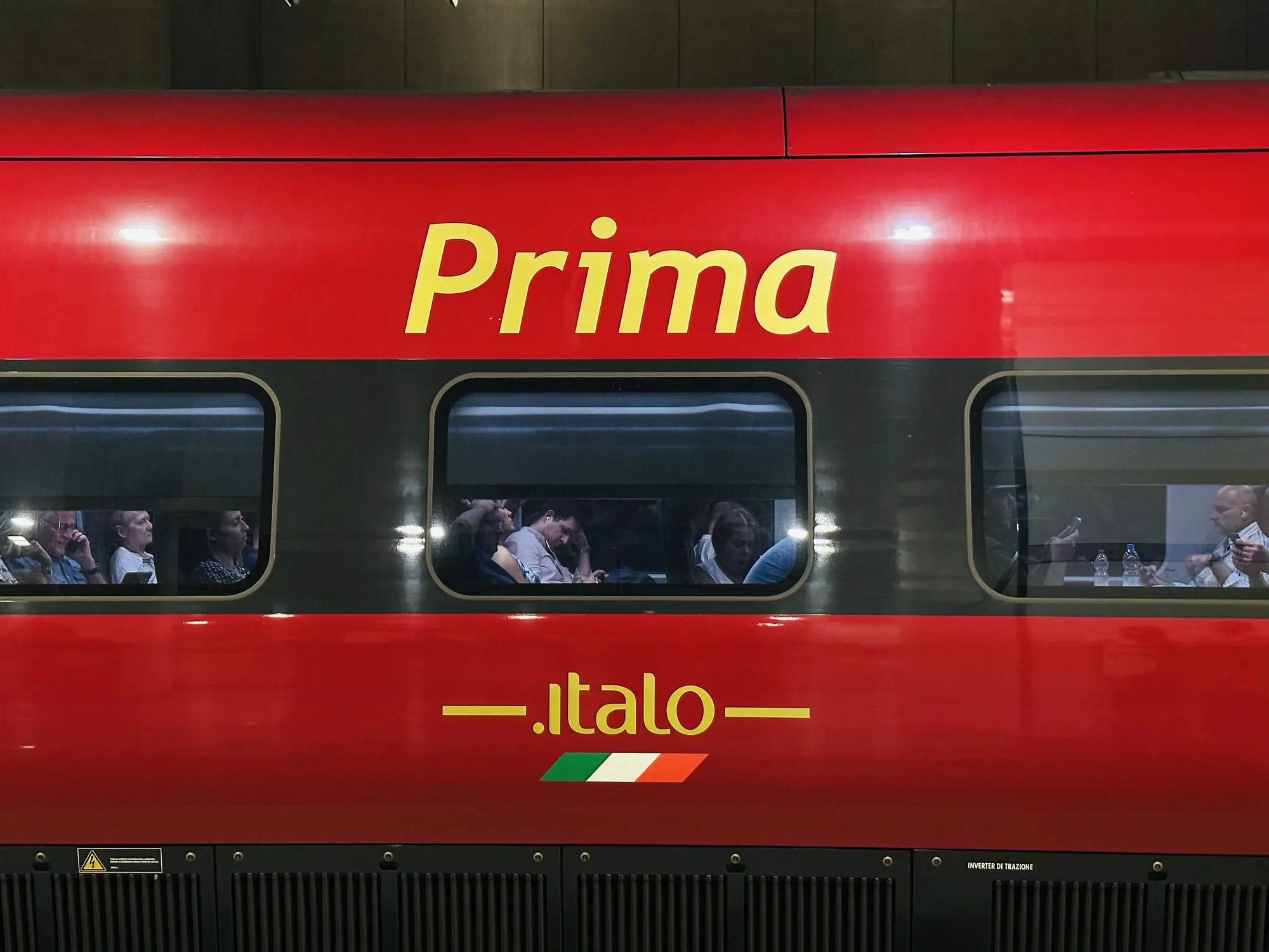 写真は、イタリアのフィレンツェで撮影された、高速鉄道イタロの「Prima」クラスの車両の外観を捉えています。鮮やかな赤色の車体に、黄色で「Prima」と大きく書かれており、その下に、イタロのロゴとイタリア国旗のカラーラインがデザインされています。窓からは、乗客の様子がうかがえ、スマートフォンを操作したり、談笑したり、飲み物を飲んだりしている様子が見られます。車両の下部は黒色のパネルで覆われており、機械的な構造が感じられます。全体的に、洗練されたモダンなデザインと、快適な移動空間が組み合わさった印象を与えます。