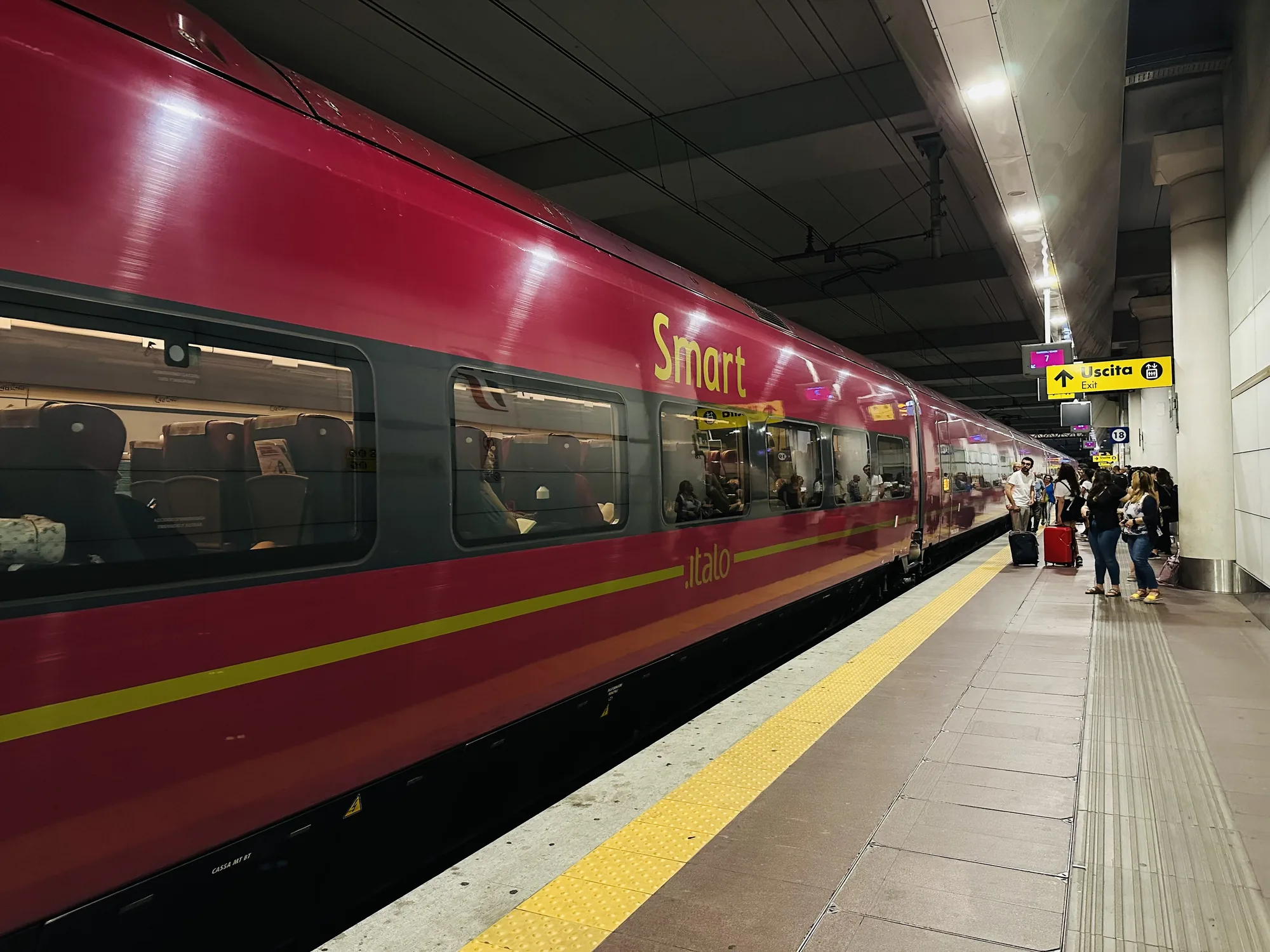 写真はイタリアのフィレンツェにある駅のホームで撮影されたもので、赤色のイタロ（Italo）高速鉄道が写っています。列車は「Smart」クラスで、窓からは乗客の姿が見えます。ホームには乗客と思われる人々が何人かいて、荷物を持っていたり、列車の到着を待っている様子です。駅の天井には照明と配線が張り巡らされており、ホームの床には黄色い点字ブロックが敷かれています。看板には出口（Uscita/Exit）の案内が表示されています。全体的に、駅の活気と列車の旅の雰囲気が伝わってきます。