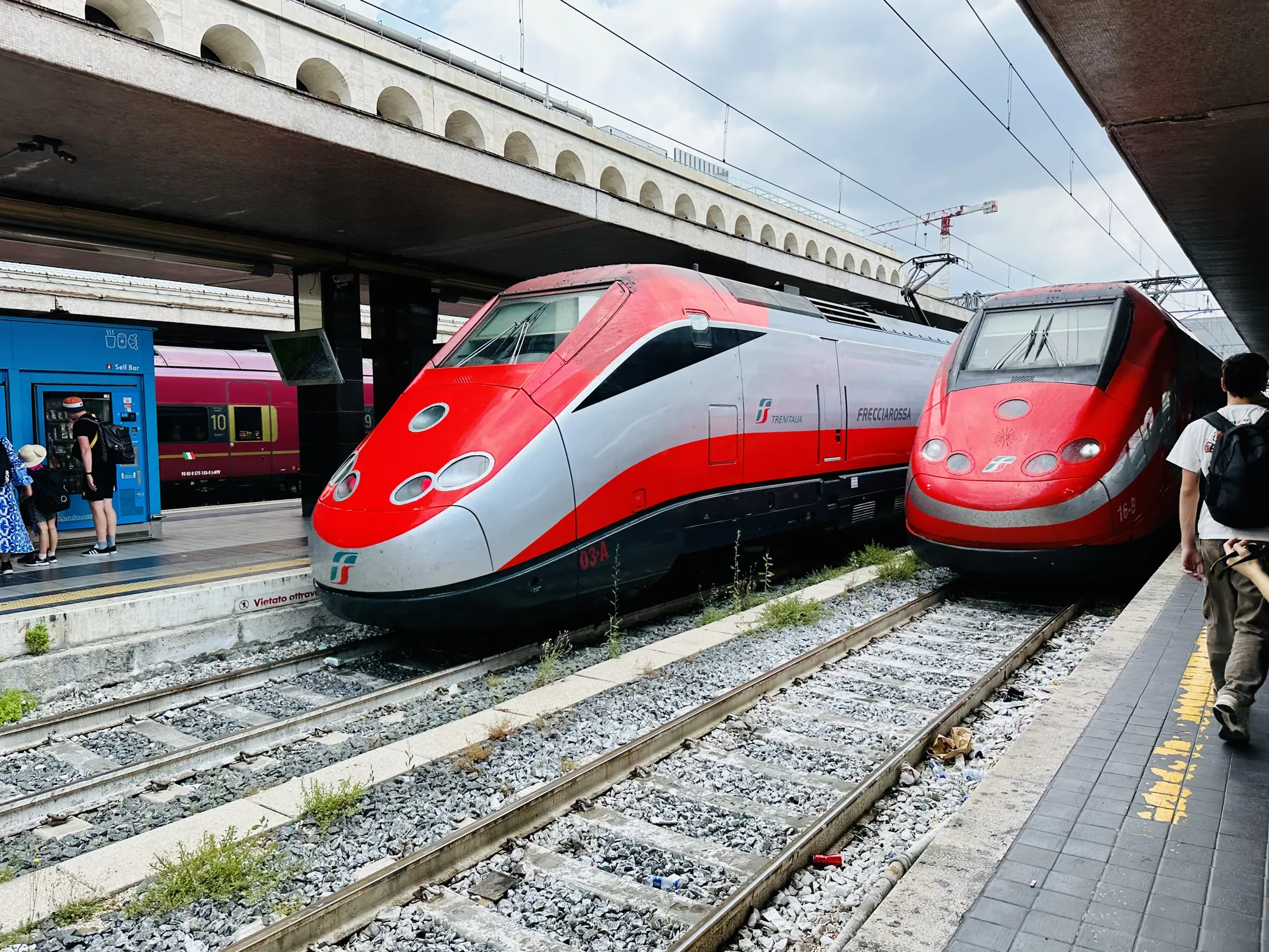 イタリアのフィレンツェの駅で撮影された写真です。赤とグレーの高速列車フレッチャロッサが2編成並んで停車しています。ホームには人がいて、自動販売機のようなものがあります。背景には駅の屋根が見え、空は少し曇っています。全体的に、駅の活気ある雰囲気と、列車の近代的なデザインが印象的な写真です。線路には草が生えていたり、ごみが落ちていたりして少し生活感も感じられます。