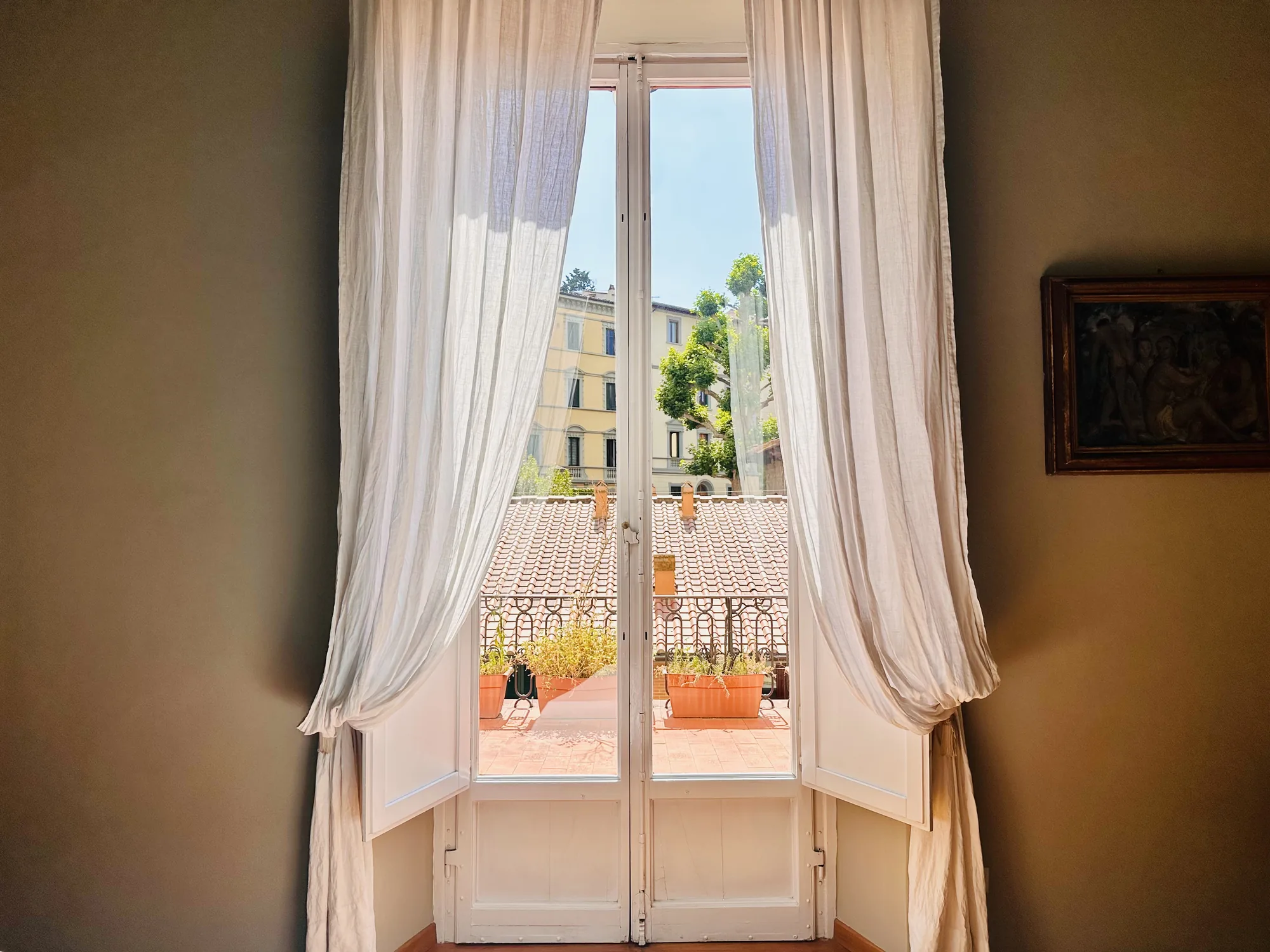 イタリア、フィレンツェで撮影されたと思われる写真です。室内の落ち着いた色合いの壁に飾られた絵画が、歴史を感じさせます。開かれた白い窓からは、テラコッタタイルのテラスが見え、植木鉢に植えられた植物が置かれています。テラスからは、フィレンツェの街並みが垣間見え、建物や木々の緑が色彩を添えています。窓にかかる薄手のカーテンが、柔らかな光を室内に取り込んでいます。全体的に、穏やかで静かな時間が流れる、フィレンツェの一室の風景を切り取った写真です。