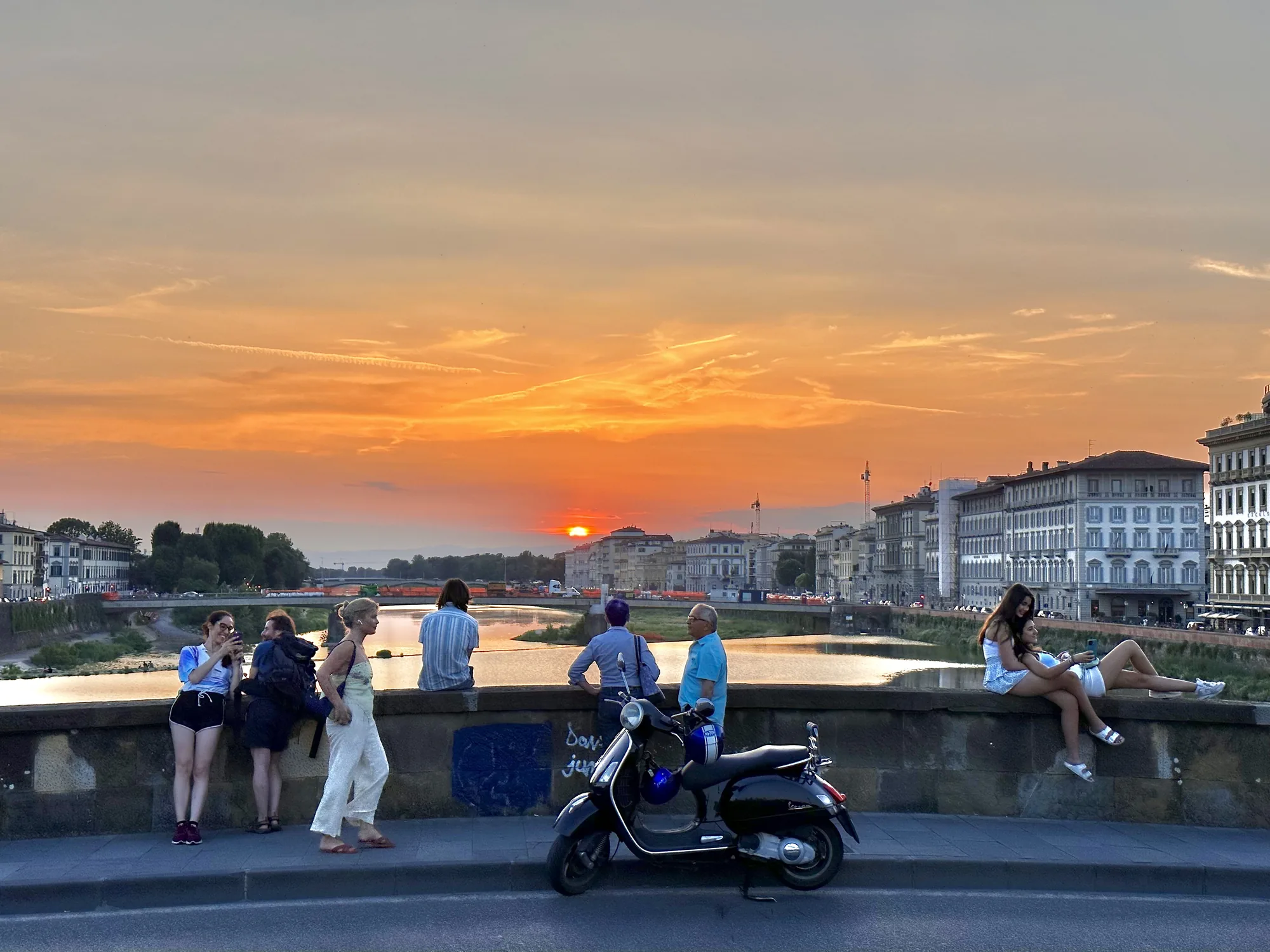イタリア、フィレンツェで撮影された夕暮れ時の写真です。夕焼け空がオレンジ色に染まり、穏やかな川面を照らしています。橋の上には数人の人々がおり、夕日を眺めたり、写真を撮ったりして、それぞれの時間を過ごしている様子です。左側には若い女性たちが立って会話をしています。中央には黒いベスパのスクーターが停められており、橋の欄干に寄りかかっている人もいます。右側には2人の若い女性が欄干に腰掛けてリラックスしています。背景にはフィレンツェの歴史的な建物群が並び、美しい景観を形作っています。全体的に穏やかでノスタルジックな雰囲気が漂う写真です。