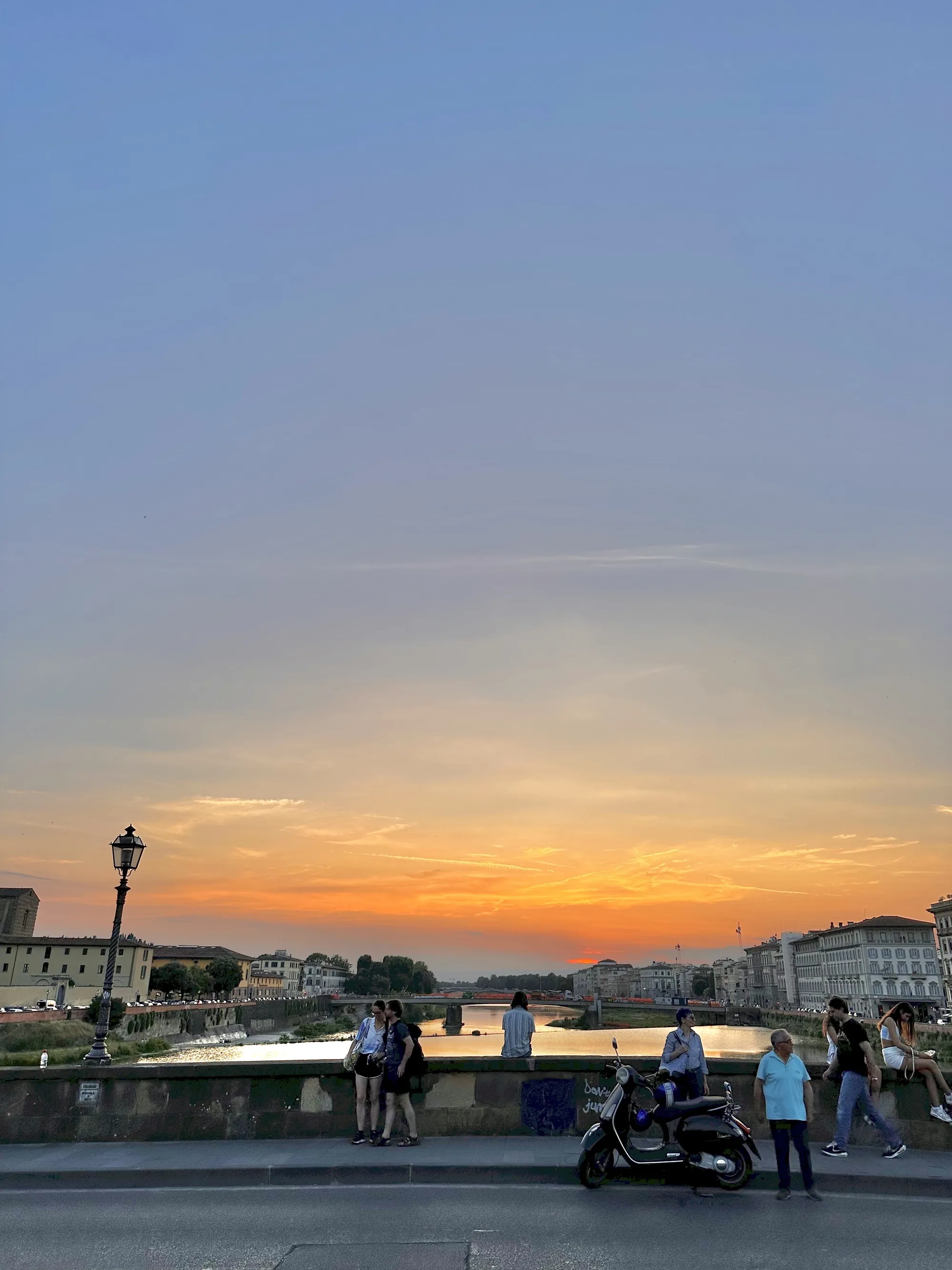 イタリア、フィレンツェで撮影された写真です。空は上部が淡い青色で、地平線に向かってオレンジ色のグラデーションに染まり、美しい夕焼けが広がっています。アルノ川が画面中央を横切り、水面には夕焼けの色が反射して輝いています。川には複数の橋が架かっており、遠景にはフィレンツェの歴史的な建物群がシルエットのように浮かび上がっています。  手前の橋の上には、人々が夕暮れの景色を楽しんでいます。数人のグループやカップルが立ち止まって景色を眺めたり、おしゃべりを楽しんだりしています。手前には黒いスクーターが停まっており、イタリアらしい風景を演出しています。写真全体は穏やかで温かい雰囲気に包まれており、フィレンツェの美しい夕暮れ時を切り取った一枚です。