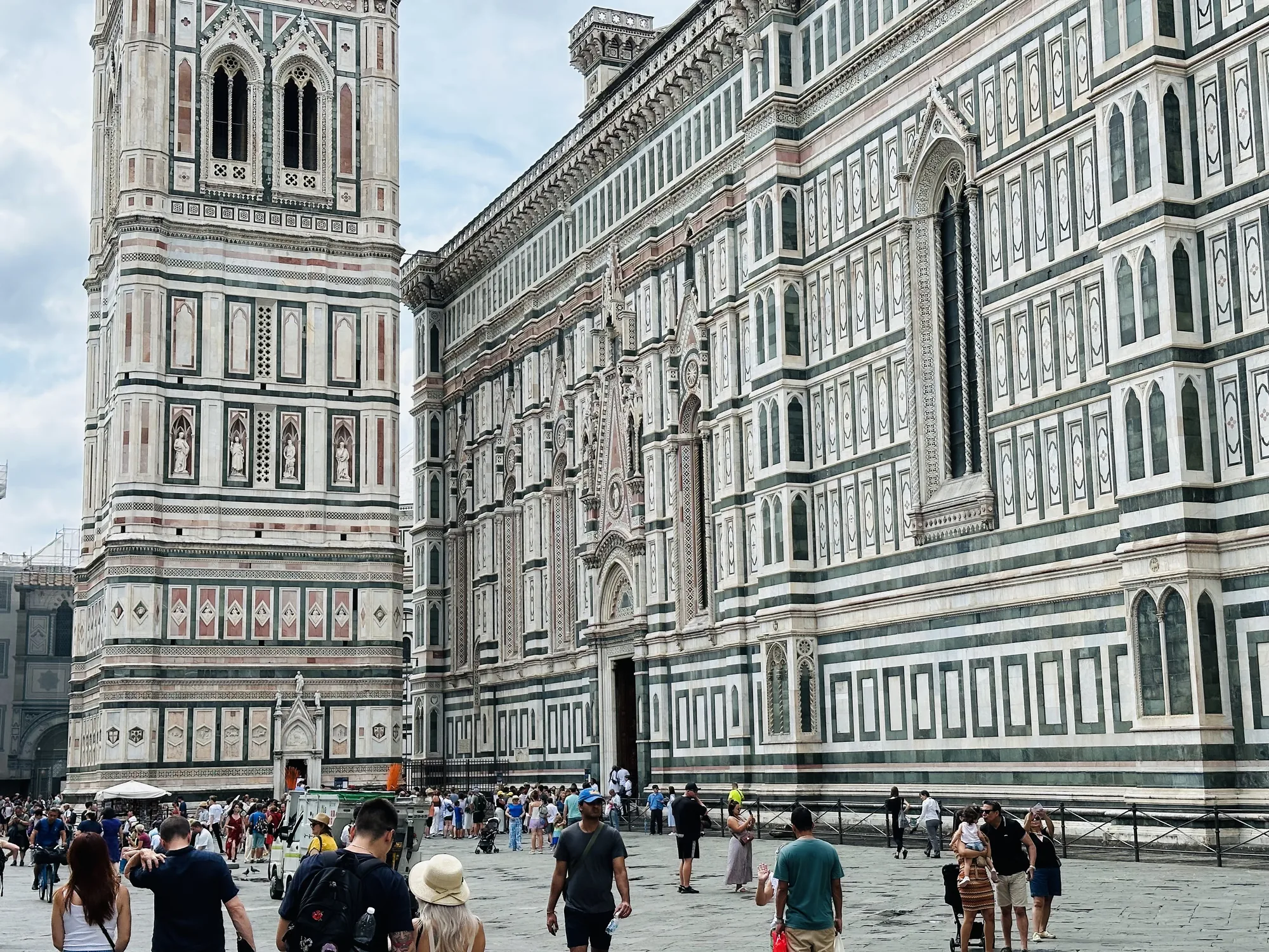 写真はイタリアのフィレンツェで撮影されたもので、フィレンツェ大聖堂（ドゥオーモ）とその周辺の様子を捉えています。手前には多くの観光客が行き交い、活気ある雰囲気が伝わってきます。大聖堂の荘厳な外観が際立っており、白、緑、ピンクの大理石を使用した複雑な装飾やゴシック様式の窓が印象的です。ジョットの鐘楼も写っており、同様の装飾が施されています。空は少し曇っており、全体的に落ち着いた色調ですが、建物の精巧な造りが細部まで見て取れます。多くの観光客がこの美しい景色を楽しんでいる様子が伺え、フィレンツェの観光名所としての人気がわかります。