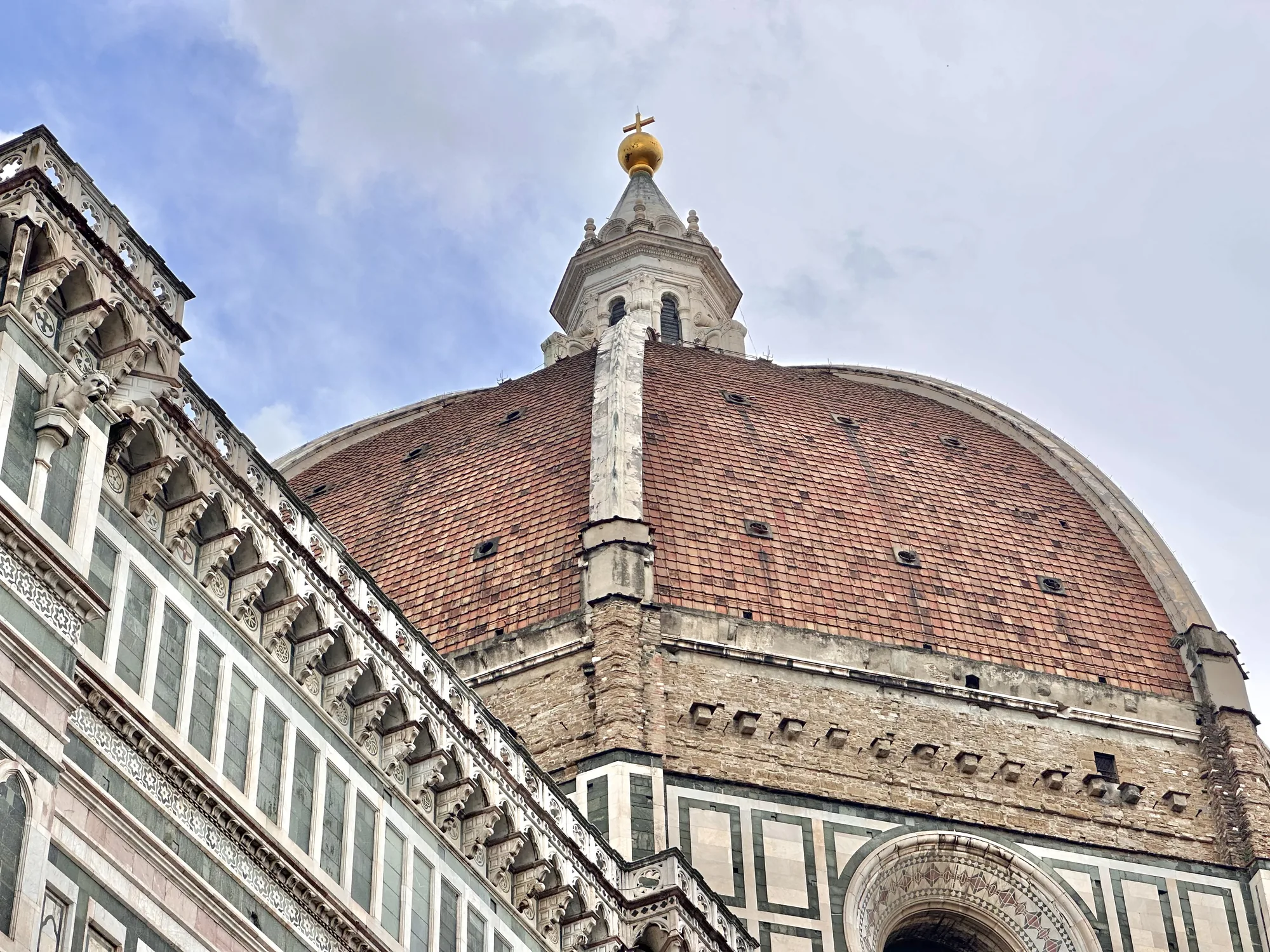 この写真は、イタリアのフィレンツェにあるフィレンツェ大聖堂 (サンタ・マリア・デル・フィオーレ大聖堂) のドームの一部を捉えたものです。ローアングルからドームを見上げて撮影されており、ドームの荘厳さと壮大さが強調されています。  ドームは、赤茶色のテラコッタの屋根瓦で覆われており、その表面は経年変化による色むらや風合いが見られます。ドームの頂上には、金色の球体と十字架が輝いており、空に向かってそびえ立っています。  ドームの基部には、白と緑の大理石を使った美しい装飾が施されており、幾何学模様やアーチ型の窓などが特徴的です。ゴシック様式の装飾も細部にわたり施されており、壮麗な雰囲気を醸し出しています。  空は曇りがちで、薄い雲が広がっており、建物全体がやや落ち着いた印象を与えています。しかし、ドームの圧倒的な存在感と、細部にまでこだわった建築様式が、見る者を魅了します。写真は、フィレンツェの象徴的な建築物である大聖堂の美しさを際立たせています。