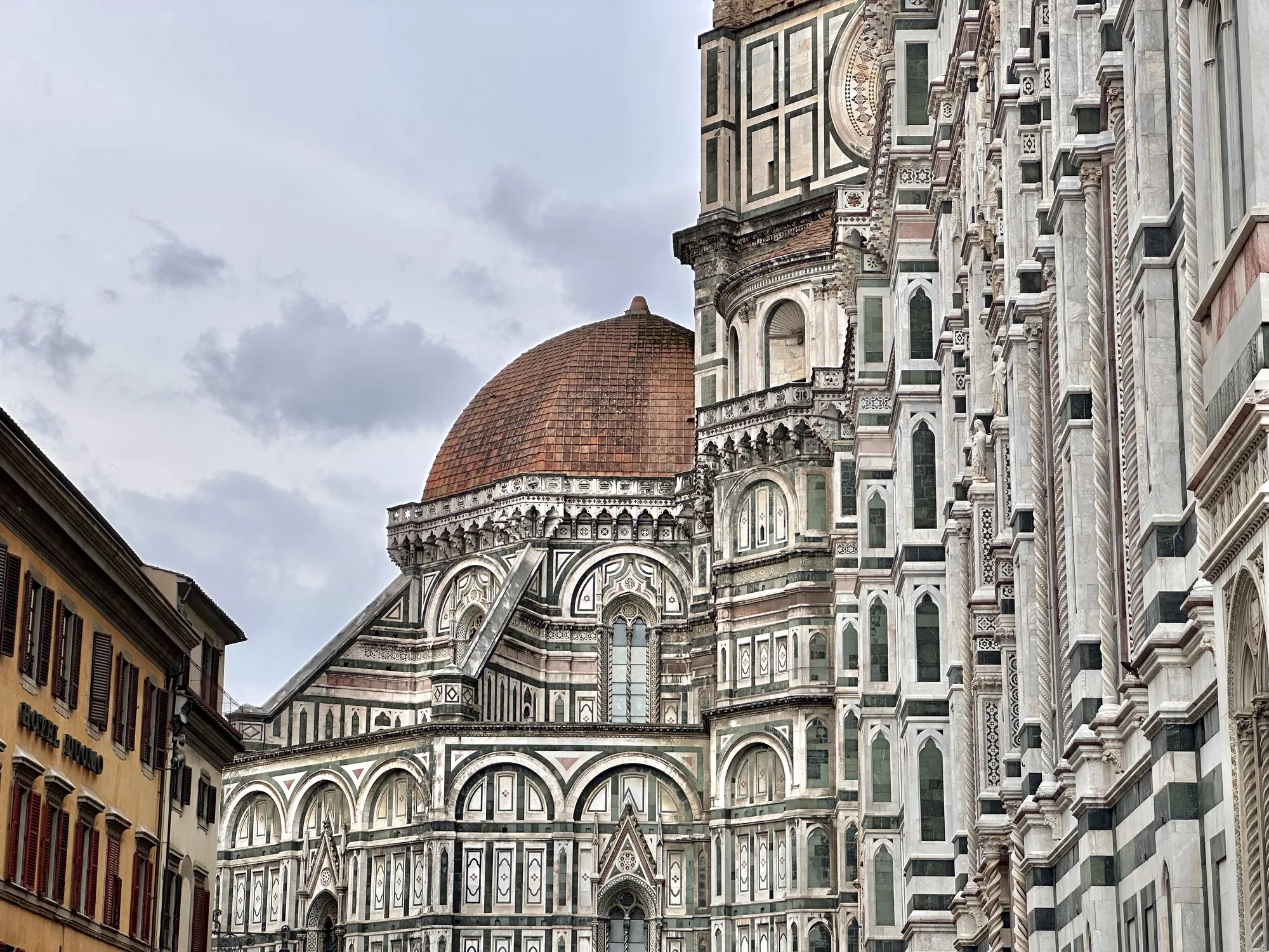 イタリアのフィレンツェで撮影された写真です。画面の中心から右側にかけて、壮麗なフィレンツェ大聖堂（サンタ・マリア・デル・フィオーレ大聖堂）の外観が大きく写っています。特に、特徴的なドームとその周辺の精緻な装飾が目を引きます。ドームは赤茶色のレンガで覆われており、歴史を感じさせる風合いです。大聖堂の壁面は、白、緑、ピンクの大理石が組み合わされ、幾何学的な模様が施されており、その壮麗さと美しさを際立たせています。ゴシック様式のアーチや窓が整然と並び、細部にわたる装飾が建物の歴史と芸術性を示唆しています。  画面の左端には、黄色がかった建物の壁面が少し写っており、窓には赤いブラインドが取り付けられています。この建物は、おそらく大聖堂に隣接する他の建造物の一部であり、背景の空は灰色がかっており、曇りの天候であることが伺えます。写真全体は、フィレンツェの歴史的な景観と建築物の美しさを捉えており、観光名所としての魅力を伝えています。