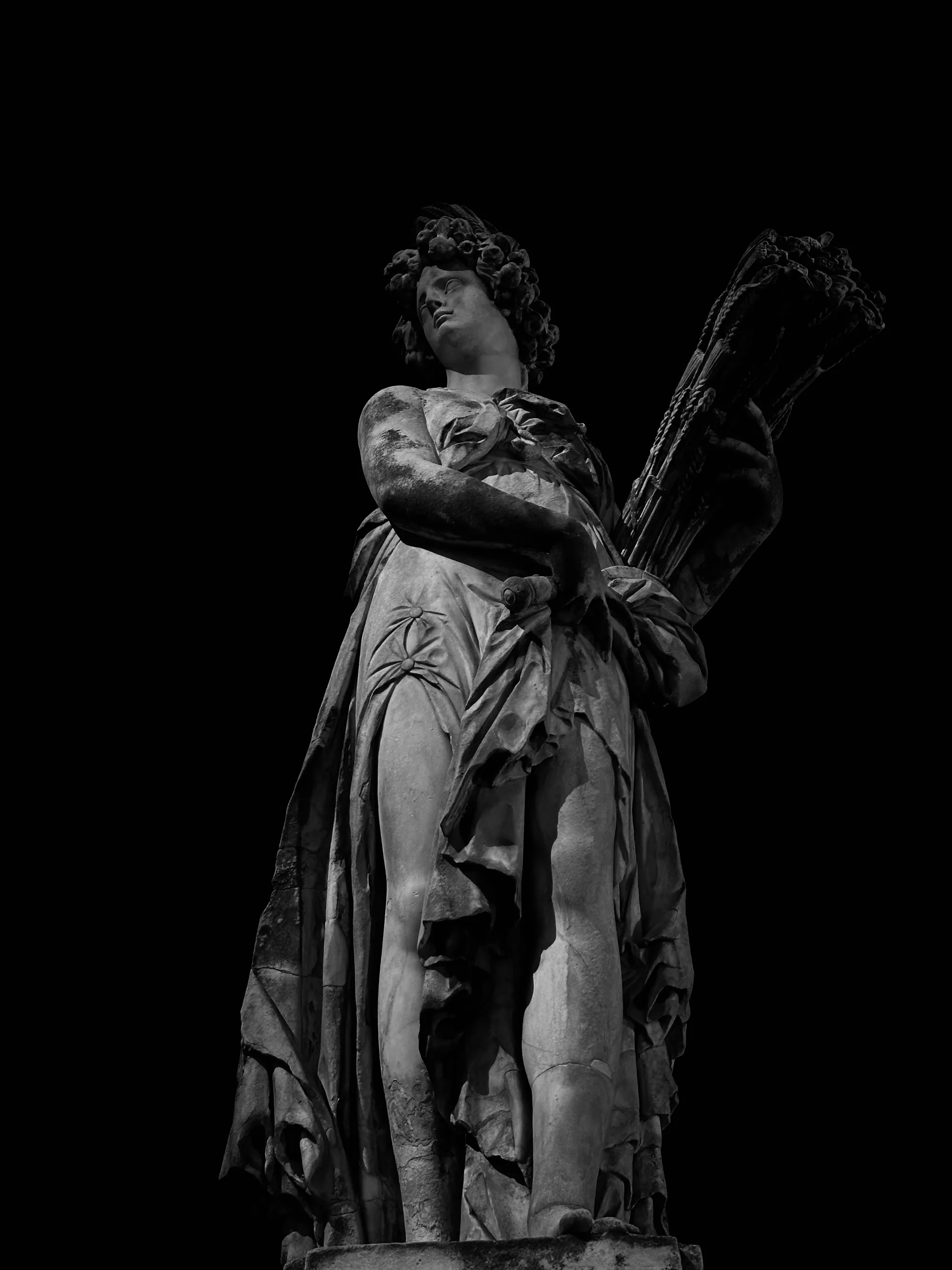 イタリアのフィレンツェで撮影された、モノクロ写真です。画像には、下から見上げる形で女性の像が写っています。像は古代ギリシャまたはローマの女神を思わせる姿で、巻き毛の髪に頭飾りをつけ、古代風の衣装を身に着けています。衣装は肩から流れるようにドレープしており、片方の脚が部分的に露出しています。像は片手で麦束のようなものを抱えています。背景は黒で塗りつぶされているため、像のディテールが強調されています。像の表面は経年劣化しており、風化や損傷が見られます。