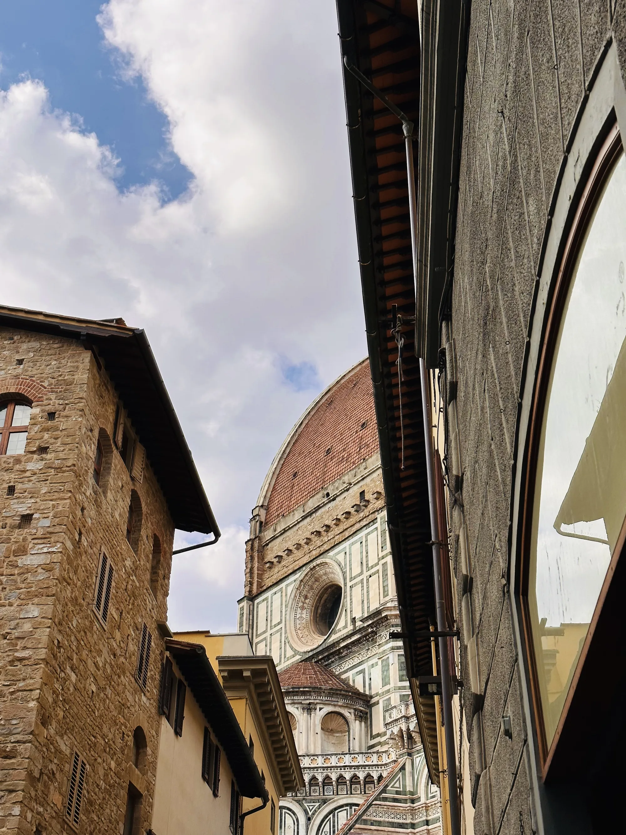 イタリアのフィレンツェで撮影された写真です。狭い路地の両側に建つ建物越しに、壮大なフィレンツェのドゥオーモ（サンタ・マリア・デル・フィオーレ大聖堂）のドームが印象的にそびえ立っています。空は青空が広がり、白い雲が点在しています。左側の建物は、石造りの壁と小さな窓が特徴的な歴史を感じさせる佇まいです。右側の建物は、グレーの壁にアーチ型の窓があり、窓には周囲の景色が映り込んでいます。写真全体に、フィレンツェの歴史的な美しさと建築の素晴らしさが凝縮されており、訪れる人々を魅了する街の魅力を伝えています。
