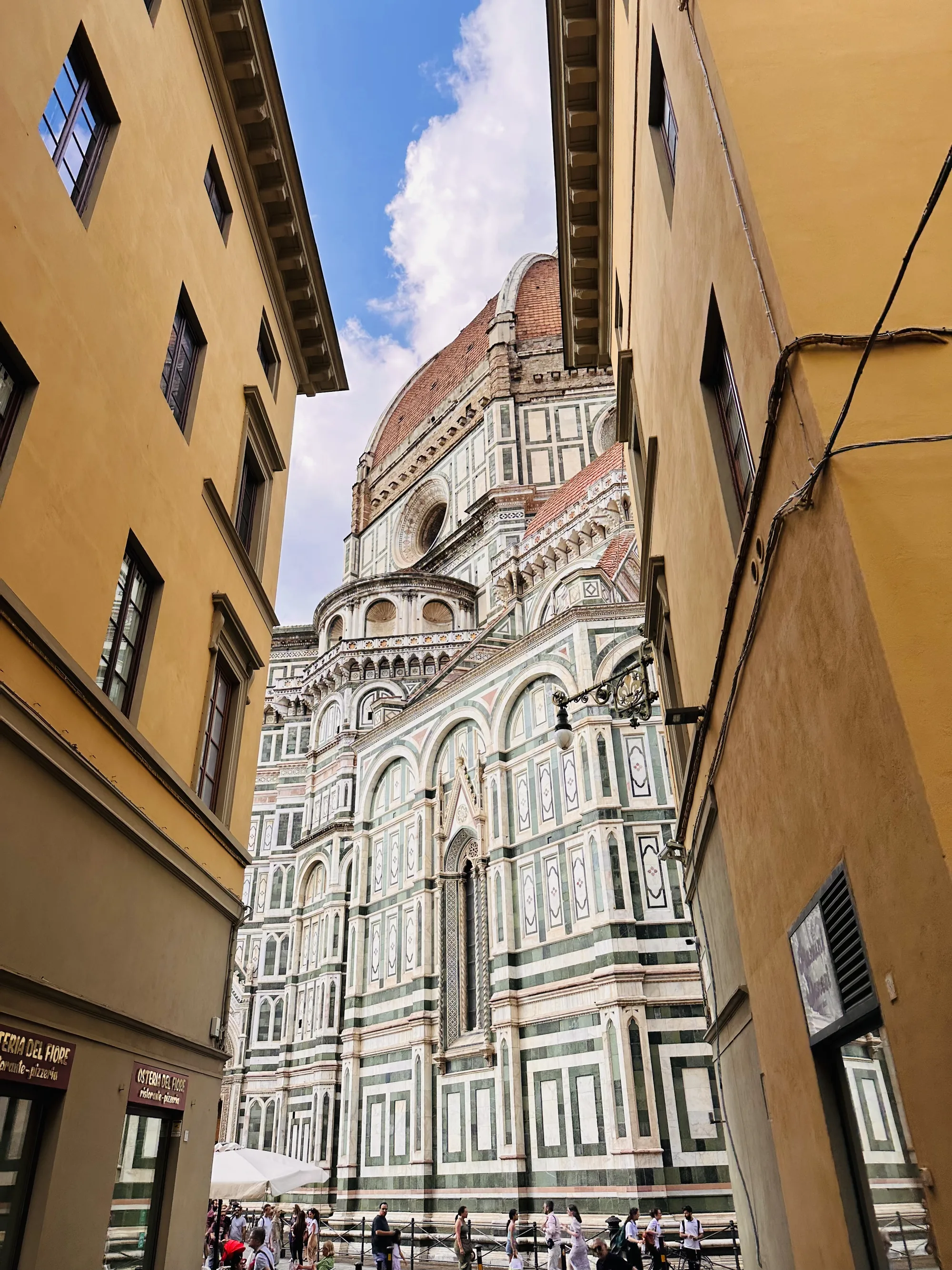 この写真はイタリアのフィレンツェで撮影されたものです。狭い路地の間から、壮大なフィレンツェのドゥオーモ（サンタ・マリア・デル・フィオーレ大聖堂）が仰ぎ見えます。ドゥオーモの独特なレンガ色のクーポラが青空に映え、その美しい外観は白と緑の幾何学模様で飾られています。  路地の両側には黄色い壁の建物がそびえ立ち、歴史を感じさせる窓枠や装飾が見られます。地面には人々が歩いており、観光客が多いことが伺えます。左側の建物には「OSTERIA DEL FIORE ristorante-pizzeria」と書かれた看板が見え、レストランであることがわかります。 全体的に、フィレンツェの歴史的な景観と活気が伝わる写真です。