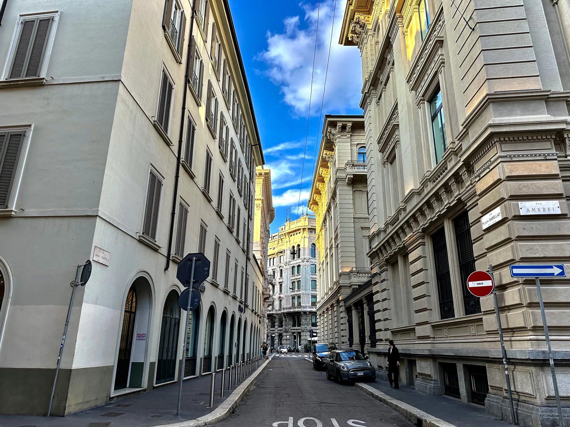 イタリア、ミラノの街中で撮影された写真です。画面中央の道路が奥へと伸び、両側には歴史を感じさせる石造りの建物が並んでいます。建物の窓にはシャッターがついており、ヨーロッパらしい風景です。空は晴れており、青空と白い雲が美しいコントラストを描いています。道路には停車している車が数台見られ、歩いている人もいます。道路には「STOP」の文字が書かれています。右手前の建物には「VIA AMEDEI」と書かれた道路標識が見えます。全体的に落ち着いた雰囲気で、ヨーロッパの街並みを堪能できる写真です。