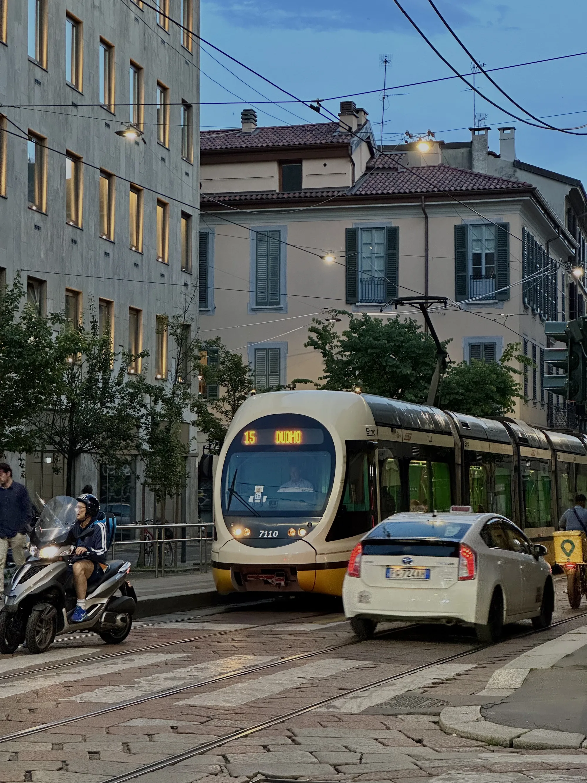 イタリアのミラノで撮影された写真です。夕暮れ時の落ち着いた雰囲気で、街の様子が写し出されています。画面中央には白いトラムが走っており、前面には「15 DUOMO」と表示されています。トラムの横には白い車が停車しており、背後には自転車に乗った人物がいます。左側には三輪バイクに乗った男性がおり、その奥には歩行者が写っています。背景には、窓に灯りがともる建物や、赤い屋根の建物が並んでいます。電線が空中に張り巡らされており、街灯が点灯しています。石畳の道路が、ヨーロッパの街並みを特徴づけています。全体的に落ち着いた色調で、静かな夕暮れ時のミラノの雰囲気が伝わってくる写真です。
