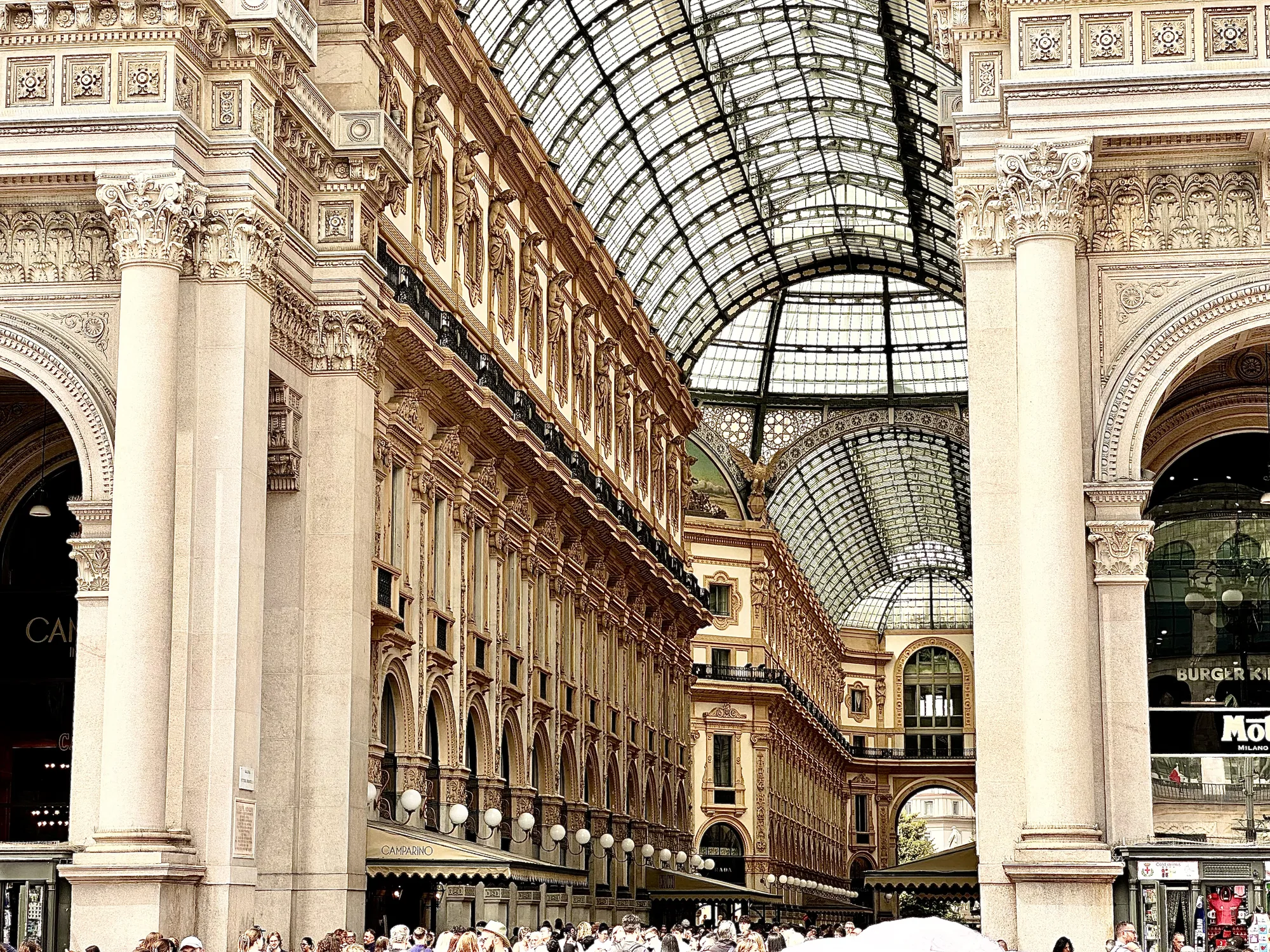 イタリア、ミラノのガレリアの内部を撮影した写真です。左右対称の構図で、壮麗な建築物が鑑賞できます。アーチ状の天井はガラス張りで、自然光が差し込み、明るく開放的な雰囲気を醸し出しています。建物の壁面はベージュ色で、緻密な彫刻や装飾が施されており、歴史と芸術を感じさせます。壁には女性の彫刻が複数飾られています。1階には店舗が並び、多くの人々で賑わっている様子が伺えます。全体的に、上品で洗練された空間が表現されています。