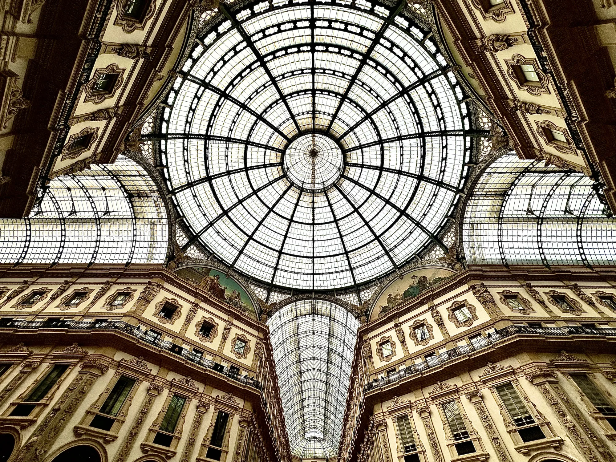 イタリアのミラノで撮影された写真です。天井を見上げた構図で、ガラス張りのドーム型の屋根が大きく写っています。屋根は格子状に組まれた金属のフレームで支えられており、太陽光が柔らかく差し込んでいます。屋根の周囲は、細かな装飾が施された建物に囲まれており、窓やバルコニーが見えます。建物の上部には絵画が飾られています。豪華で壮麗な雰囲気が感じられます。