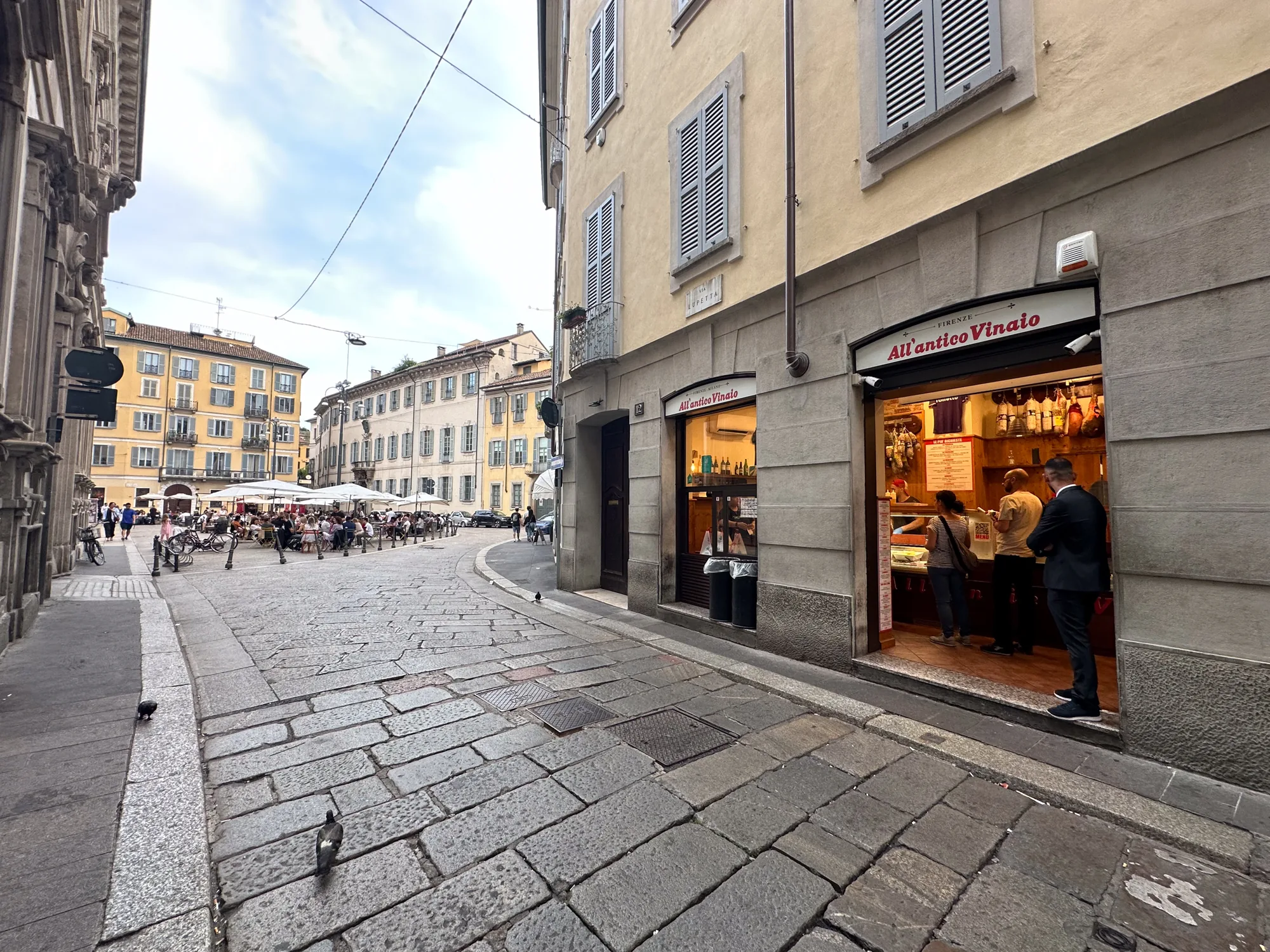 イタリアのミラノで撮影された写真です。石畳の路地が奥へと続いており、右手には飲食店「All'antico Vinaio」があります。店内には客が数人おり、賑わっている様子がうかがえます。路地の先には広場があり、パラソルの下で人々が食事を楽しんでいます。建物は伝統的なヨーロッパの建築様式で、石造りの壁や窓のシャッターが特徴的です。空は少し曇っており、落ち着いた雰囲気です。路面には鳩が2羽います。全体として、歴史的な街並みの中で人々が日常を楽しんでいる様子が伝わってきます。