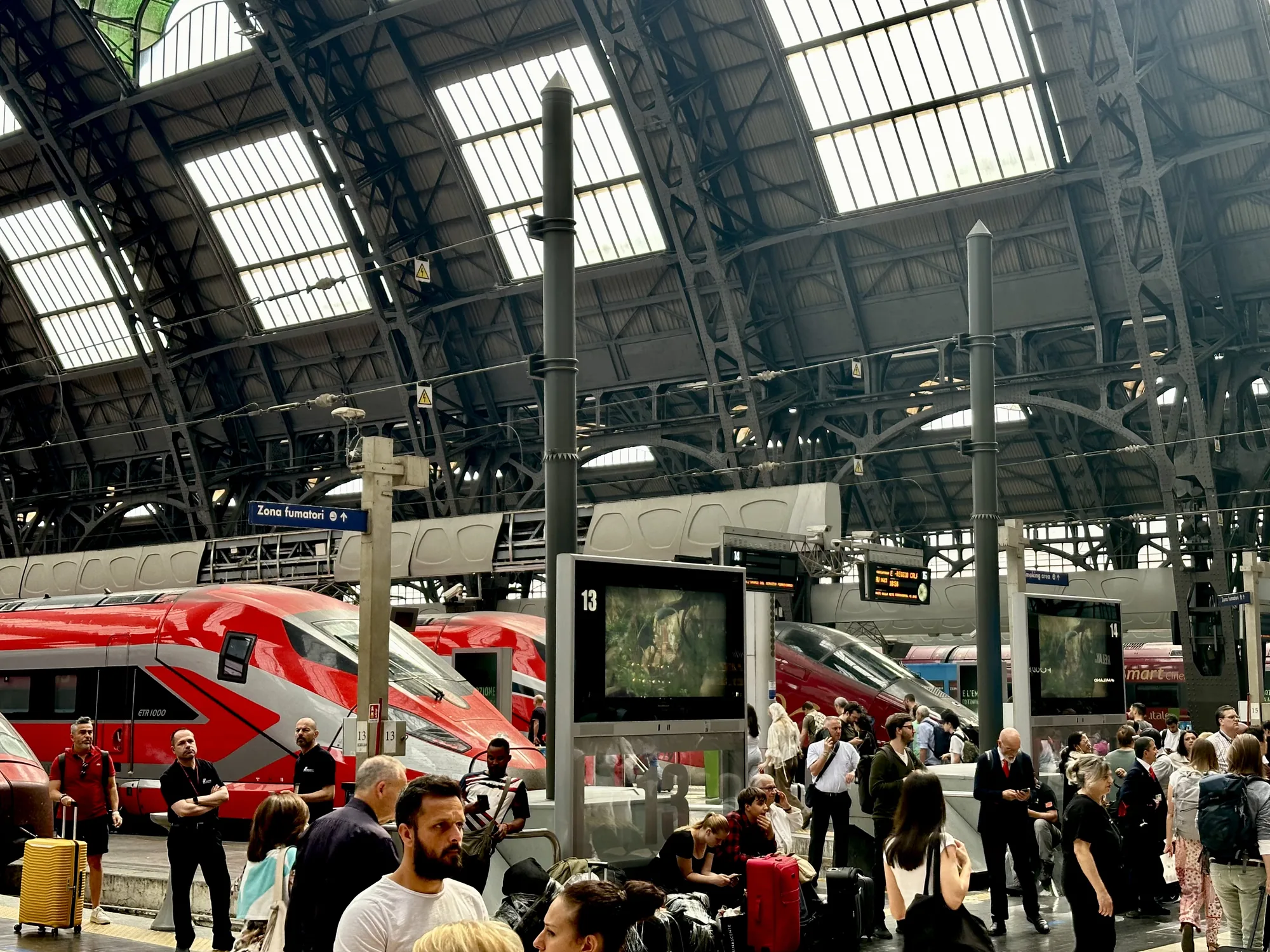 写真はイタリアのミラノ中央駅のホームで撮影されたもので、多くの人々が列車を待っている様子が写っています。背景には、赤とグレーのカラーリングが印象的な高速列車が複数停車しており、ホームには、スーツケースやバッグを持った旅行者、駅員と思われる人々が混在しています。天井は鉄骨構造で、ガラス窓から自然光が差し込み、駅構内を明るく照らしています。ホームの中央には、列車の発着情報を表示するモニターが設置されており、人々はそれを確認しながら待機しています。全体的に、駅の活気と旅の始まりの期待感を感じさせる雰囲気です。看板には「Zona fumatori (喫煙エリア)」の文字が見えます。