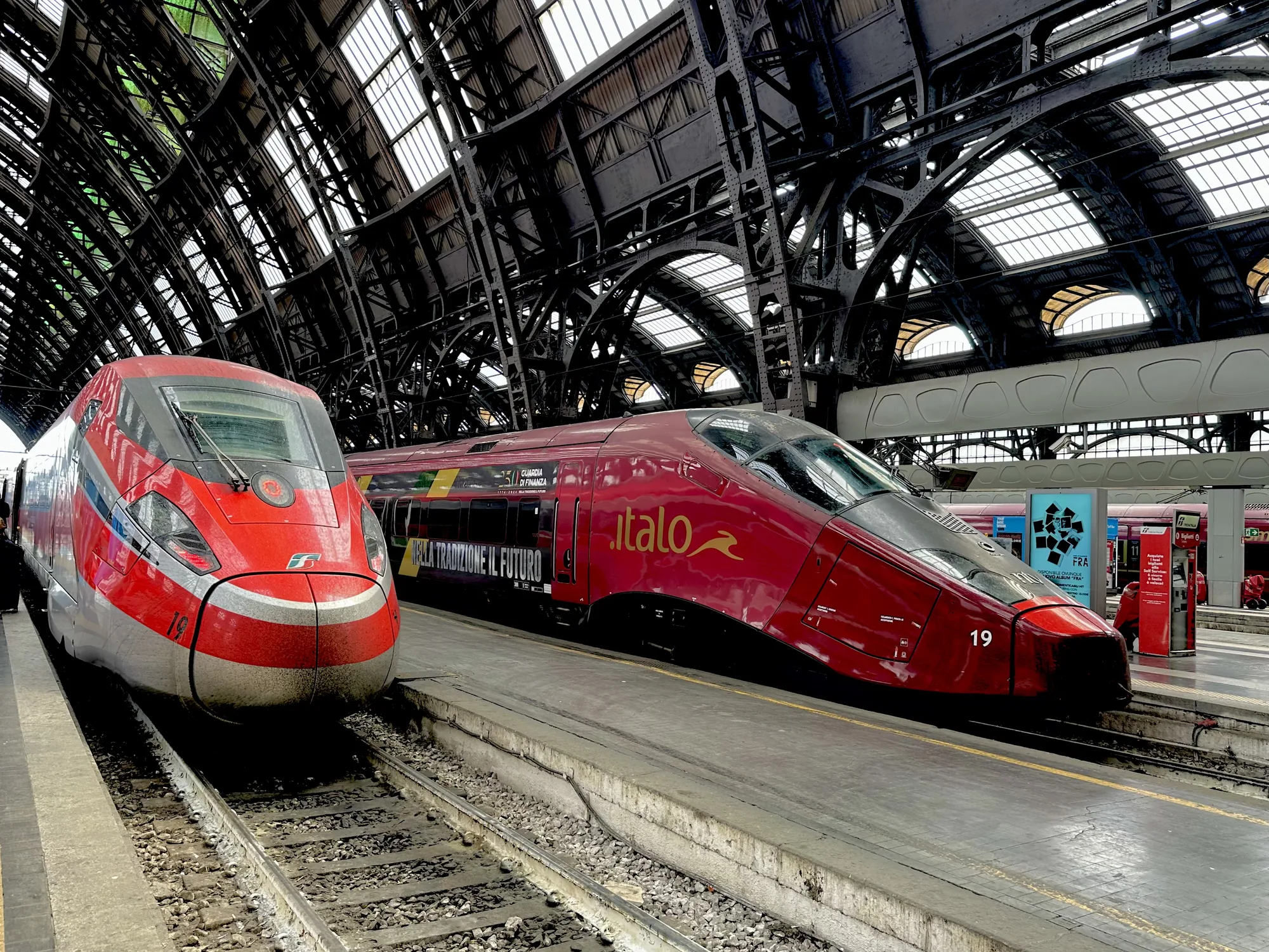 イタリアのミラノ中央駅で撮影された写真です。構内には2台の高速列車が停車しています。手前にはイタリア鉄道（Trenitalia）のフレッチャロッサ、奥にはイタロ（Italo）が写っています。両列車とも赤を基調としたカラーリングで、洗練された流線形のデザインが特徴です。イタロの車体には「NELLA TRADIZIONE IL FUTURO（伝統の中に未来がある）」というスローガンが書かれています。駅の屋根は鉄骨が複雑に組み合わされており、歴史を感じさせる重厚な雰囲気です。プラットフォームには、駅の案内板や自動券売機、広告などが設置されており、駅の賑わいを伝えています。全体的に、近代的な高速列車と歴史的な駅舎が調和した、イタリアらしい風景を捉えた写真です。