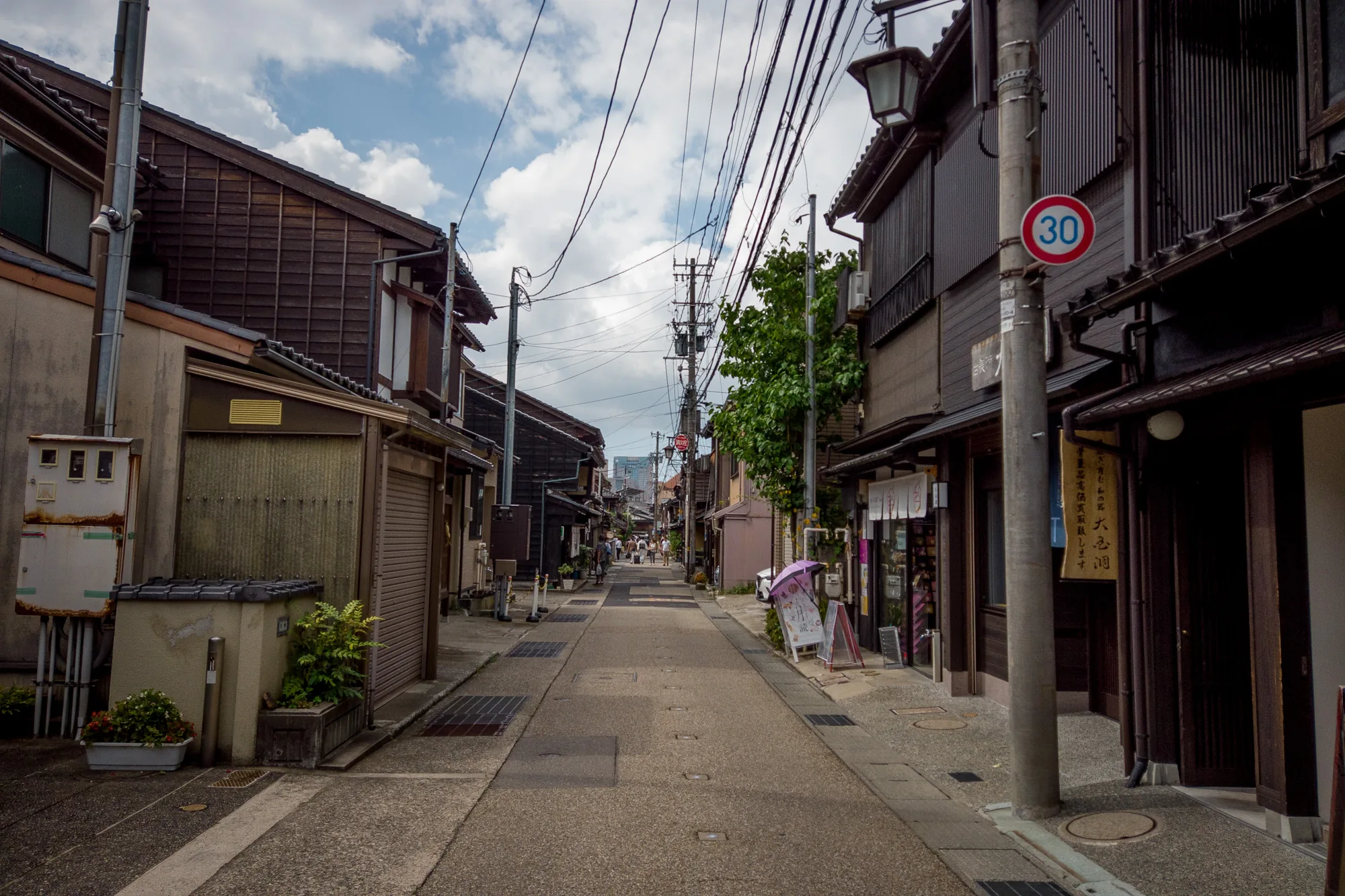 写真は石川県金沢市で撮影された、歴史的な街並みを捉えたものです。道の両側には伝統的な木造家屋が建ち並び、落ち着いた雰囲気を作り出しています。建物は濃い茶色で統一され、時代を感じさせる風格があります。空には白い雲が浮かび、電線が張り巡らされています。道路は石畳で舗装されており、整然とした印象を与えます。道の奥には、現代的な高層ビルが小さく見え、新旧の対比が面白い構図となっています。道には数人の人が歩いており、傘をさしている人もいます。路傍には植物が飾られ、生活感が感じられます。写真全体から、金沢の歴史と文化が感じられる一枚です。