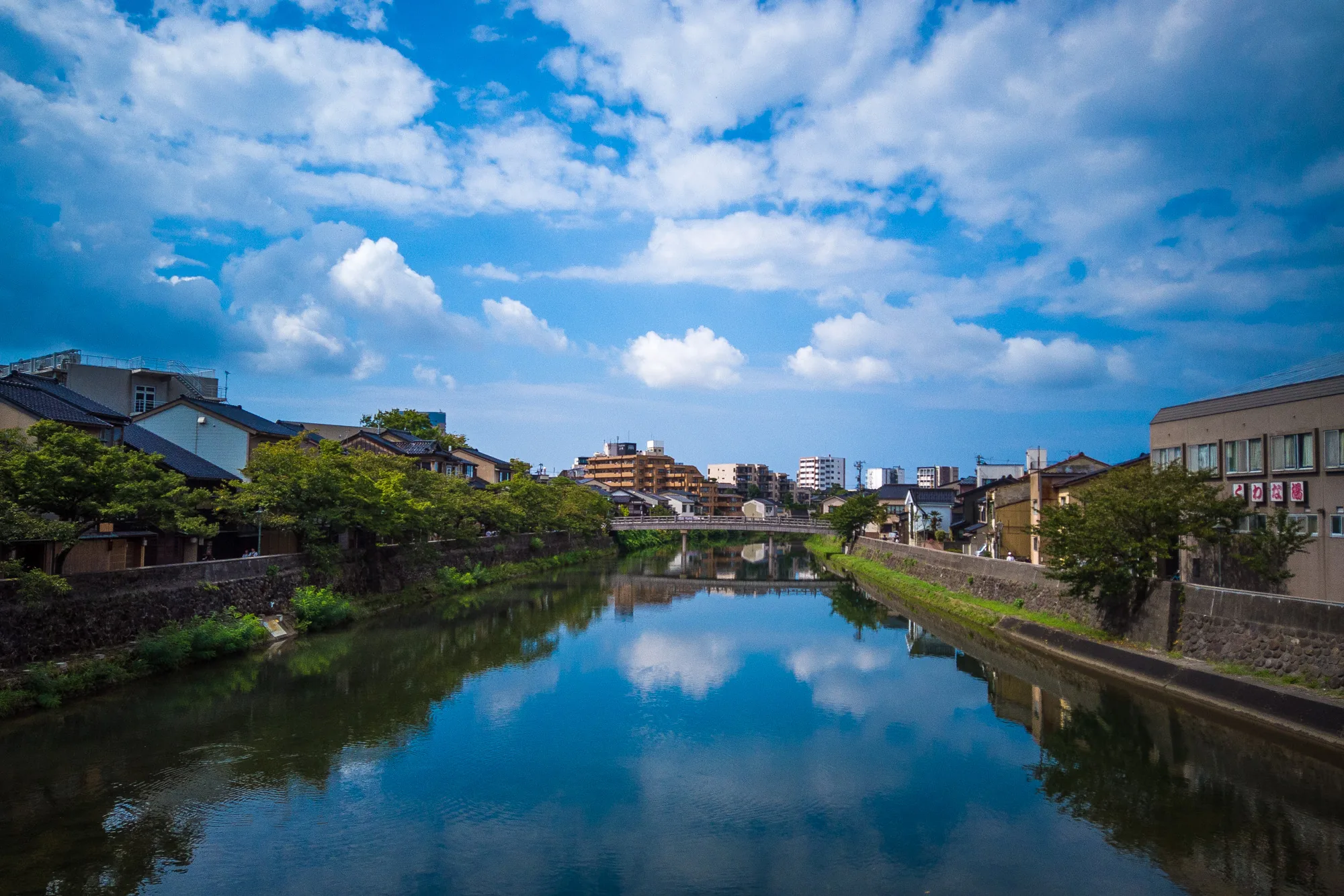 写真は日本の金沢で撮影された風景です。画面中央には、穏やかに流れる川が写っており、その水面には空の青と白い雲が鏡のように映し出されています。川の両岸には、石垣が築かれ、その上に緑の木々が生い茂っています。左岸には伝統的な日本家屋が並び、右岸にはやや近代的な建物も見られます。遠くには、高層マンションやビルが建ち並び、都市の様子を伺わせます。川には小さな橋が架かっており、その向こうにも建物が続いています。空は青く、白い雲が点在し、全体的に爽やかで穏やかな雰囲気を醸し出しています。