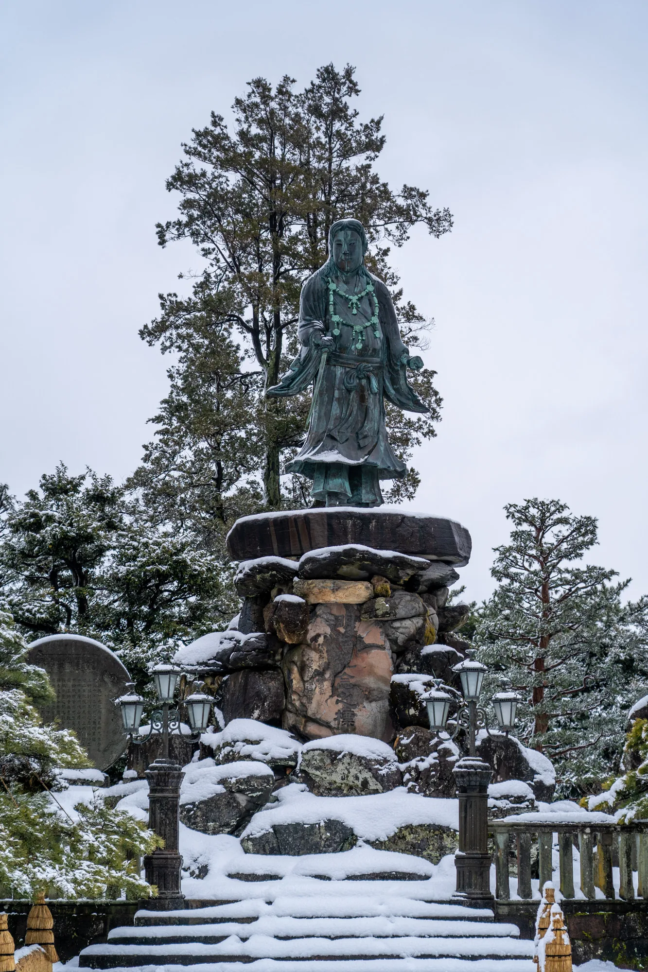 写真は、日本の金沢で撮影されたと思われる雪景色です。中央に、石積みの台座の上に立つ、青銅色の像が配置されています。像は伝統的な衣装を身につけ、首には数珠のようなものをかけています。表情は穏やかで、威厳を感じさせます。像の背景には、常緑樹がそびえ立ち、冬の空にシルエットを描いています。  像の足元には、大小さまざまな石が積み上げられ、その上に雪が積もっています。石灯籠が左右に配置され、庭園の雰囲気を醸し出しています。手前には階段があり、こちらも雪で覆われています。全体的に、静かで落ち着いた雰囲気の写真で、日本の冬の美しさを伝えています。雪の白と像の青銅色のコントラストが印象的です。庭園全体が雪化粧されており、侘び寂びの世界観を感じさせます。