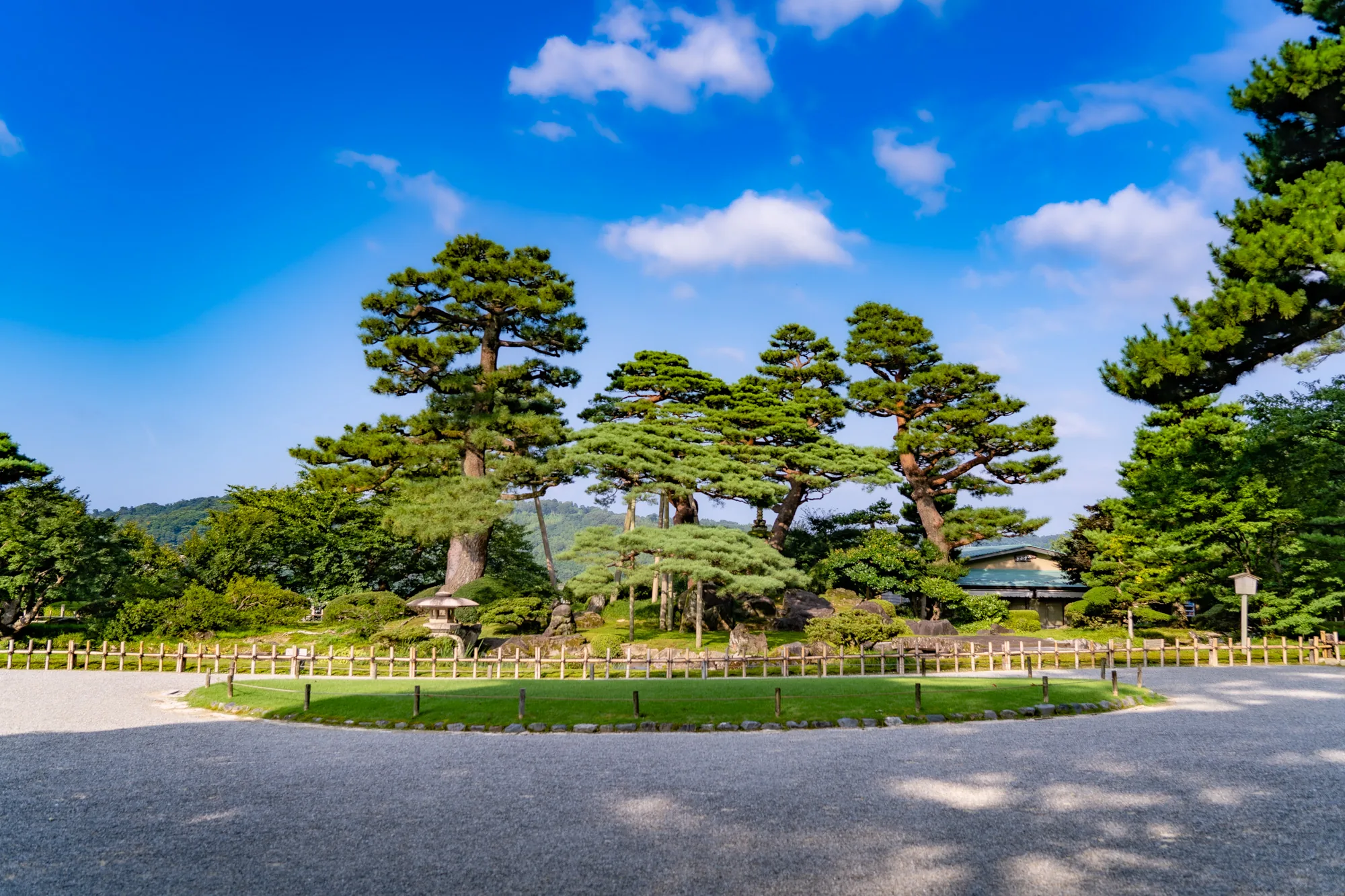 写真は、日本の金沢にある庭園の風景を捉えています。広々とした砂利道が前景を占め、その先には低い木柵で囲まれた芝生のエリアが広がっています。芝生の奥には、手入れの行き届いた庭木や岩が配置され、灯篭がアクセントになっています。背景には、堂々とした松の木々が空に向かって伸び、青い空と白い雲のコントラストが鮮やかです。庭園の奥には、屋根の見える建物があり、風景に奥行きを与えています。全体的に、静かで穏やかな日本の伝統美を感じさせる写真です。晴れた日の光が庭園全体を明るく照らし、清々しい雰囲気を醸し出しています。