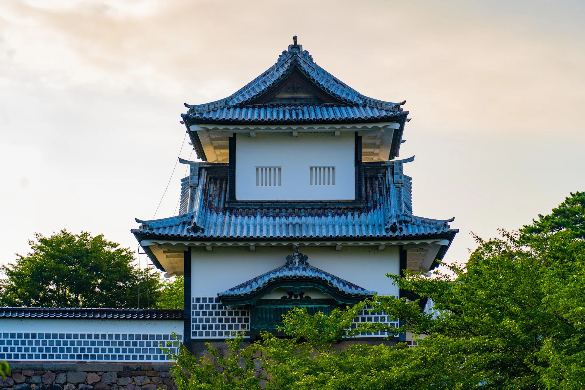 写真は日本の金沢で撮影されたもので、金沢城の櫓（やぐら）を捉えています。手前には緑豊かな木々が茂り、その奥に白壁と瓦屋根が特徴的な櫓が聳え立っています。櫓の屋根は青みがかった瓦で葺かれており、歴史を感じさせる趣があります。壁は白く塗られ、格子状のデザインが施されています。背景には薄い黄色がかった空が広がっており、夕暮れ時の穏やかな雰囲気を醸し出しています。全体的に、日本の伝統的な建築美と自然の調和が感じられる写真です。