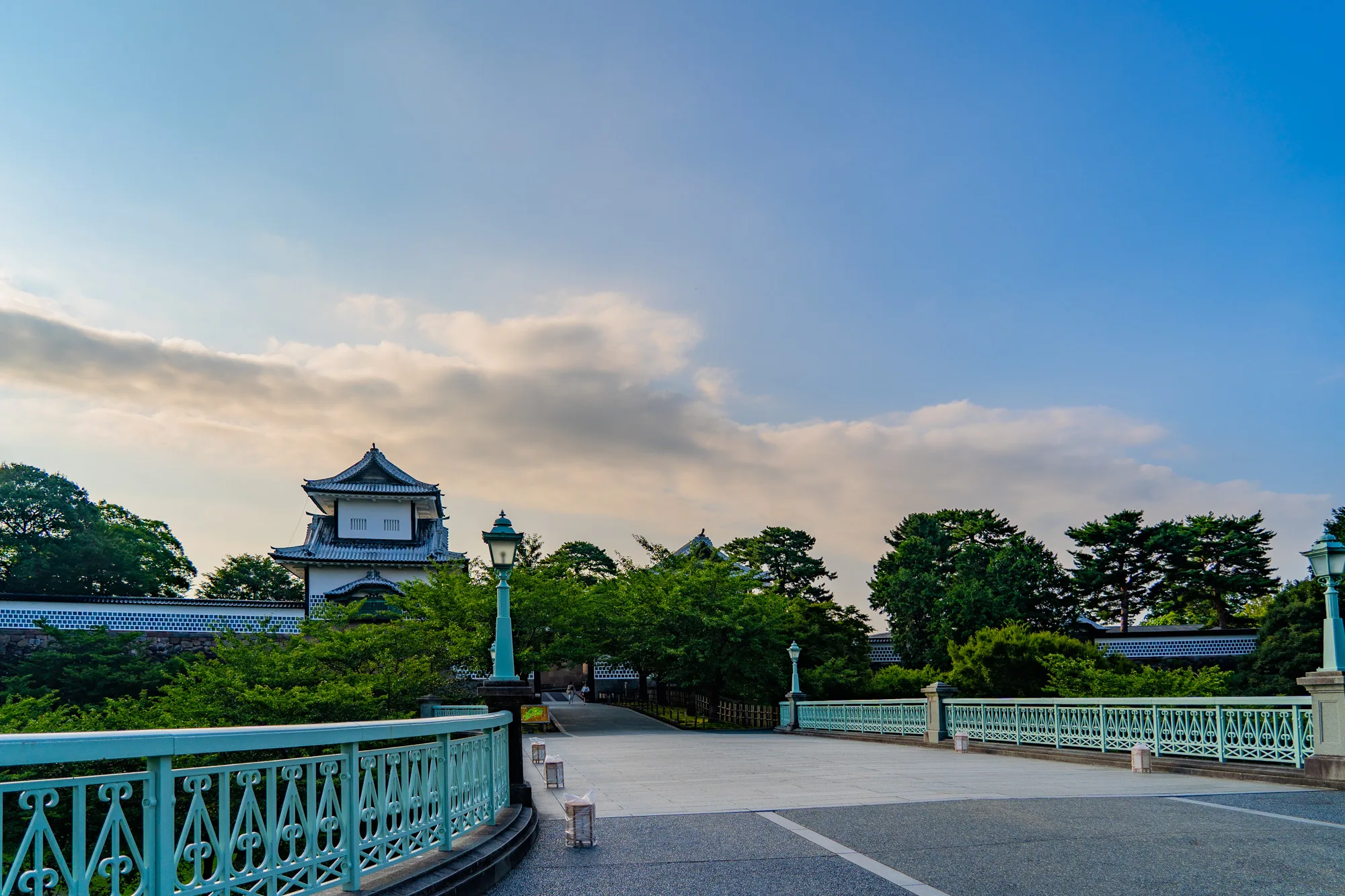 写真は、日本の金沢で撮影されたもので、金沢城へと続く橋の風景を捉えています。橋は淡い緑色の手すりが特徴で、その奥には石垣と塀に囲まれた金沢城の建物が見えます。木々の緑が豊かで、空は青く、部分的に雲が浮かんでいます。橋の上には街灯が設置されており、夕暮れ時の穏やかな雰囲気を醸し出しています。全体的に静かで落ち着いた印象の写真です。