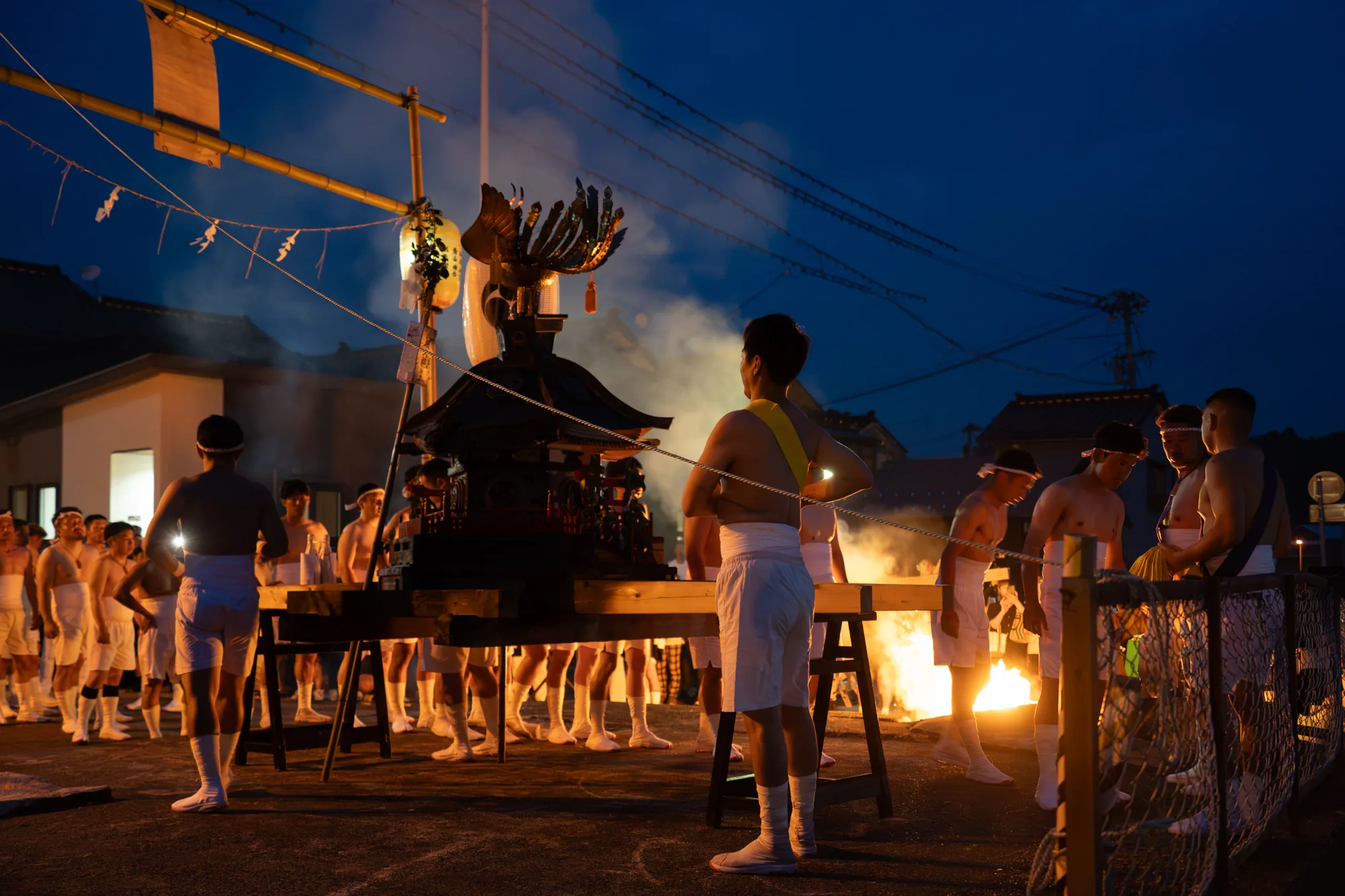 この写真は、日本の能登地方で撮影されたお祭りの様子を捉えています。時刻は夜のようで、深い青色の空が印象的です。暗闇の中、祭りの提灯や炎の光が周囲を照らし出し、幻想的な雰囲気を醸し出しています。  写真の中心には、神輿が据えられています。神輿は黒を基調とした豪華な装飾が施されており、屋根の上には金色の装飾が輝いています。神輿の周りには、白い鉢巻と白い衣装を身に着けた担ぎ手たちが集まっています。彼らは上半身裸で、祭りの熱気を帯びている様子が伝わってきます。担ぎ手の中には、黄色の襷をかけている人もいます。  写真の右側には、炎が上がっており、その熱気が伝わってくるようです。画面奥には、日本の伝統的な家屋が並んでおり、祭りの場所が日本の古い街並みであることがわかります。電線が張り巡らされているのも見えます。  全体的に、この写真は能登地方の伝統的なお祭りの力強さや熱気を表現しており、日本の文化を感じさせる一枚です。