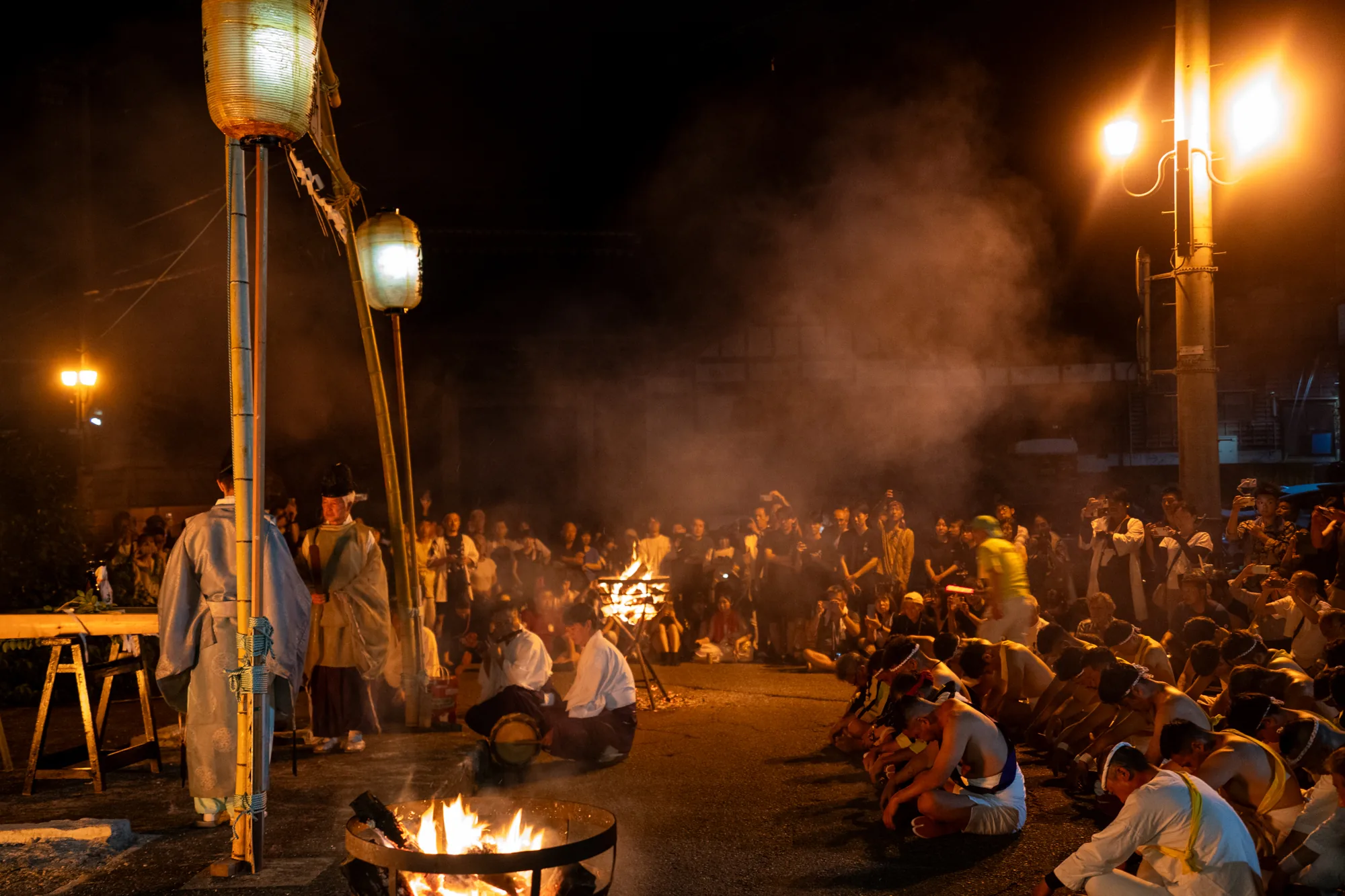 これは、日本の能登地方で撮影された夜祭りの写真です。暗闇の中、火が焚かれ、煙が立ち上っています。画面奥には大勢の人が集まっており、手前の地面には、白い鉢巻をした若者たちが整列して座っています。上半身裸の人もおり、祭りの熱気を感じさせます。左手には提灯が吊るされ、神官のような服装をした人が立っています。右手には街灯が明るく光り、その光が煙を照らし出しています。群衆はカメラやスマートフォンで写真を撮っており、この祭りの様子を記録しようとしているようです。全体的に神秘的で、伝統的なお祭りの雰囲気が伝わってきます。