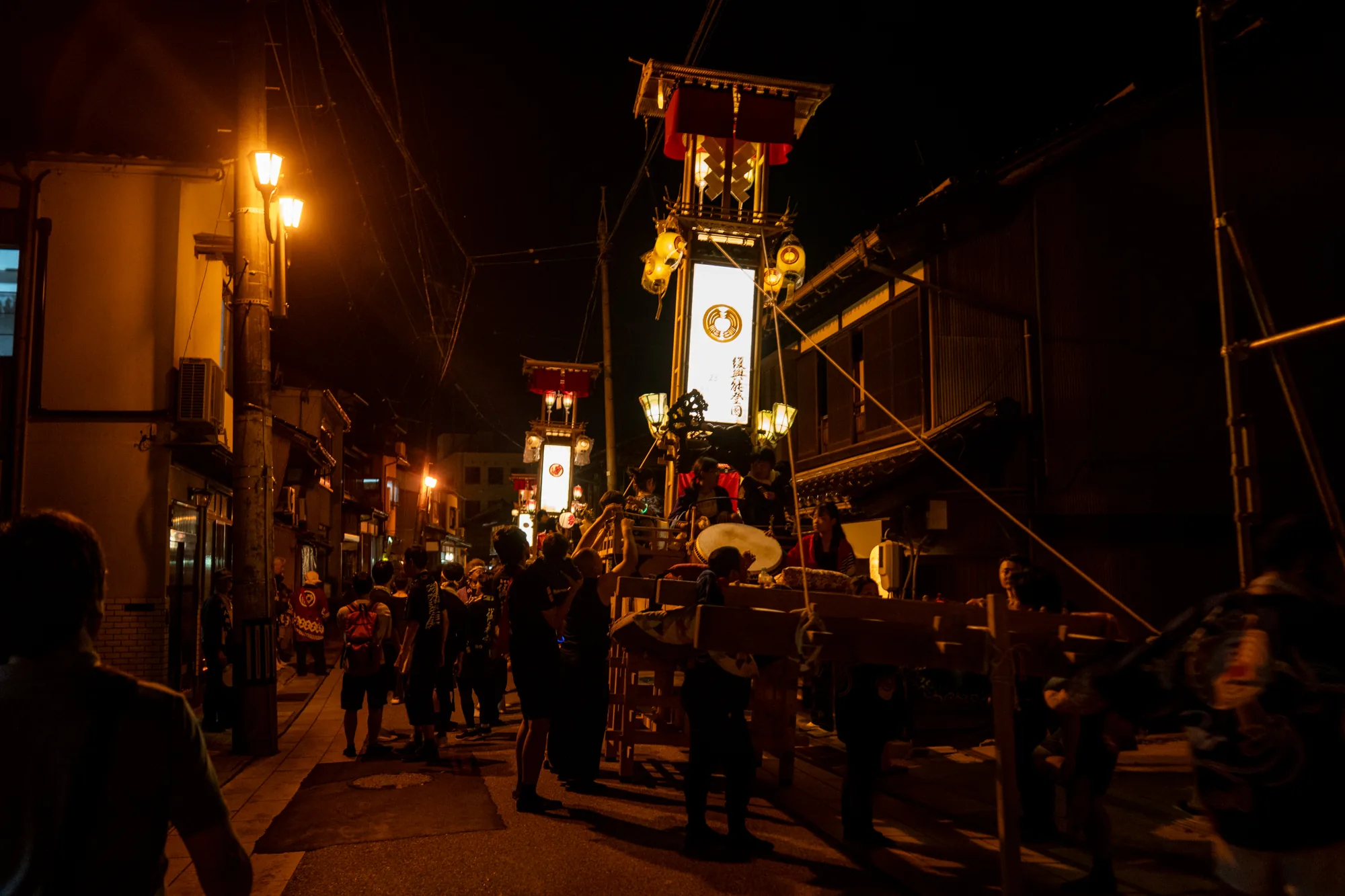写真は日本の能登で撮影された夜祭りの様子を捉えています。暗闇の中、街灯と祭りの提灯の明かりが印象的です。道の両側には古い家並みが並び、その間を大勢の人が歩いています。人々は祭り衣装を身につけ、太鼓や笛の音が聞こえてきそうです。中心には大きく装飾された祭りの山車がいくつか見え、それぞれに提灯が飾られています。山車は人々によって担がれており、祭りの熱気が伝わってきます。全体的に、伝統的な日本の祭りの賑わいと活気が感じられる写真です。