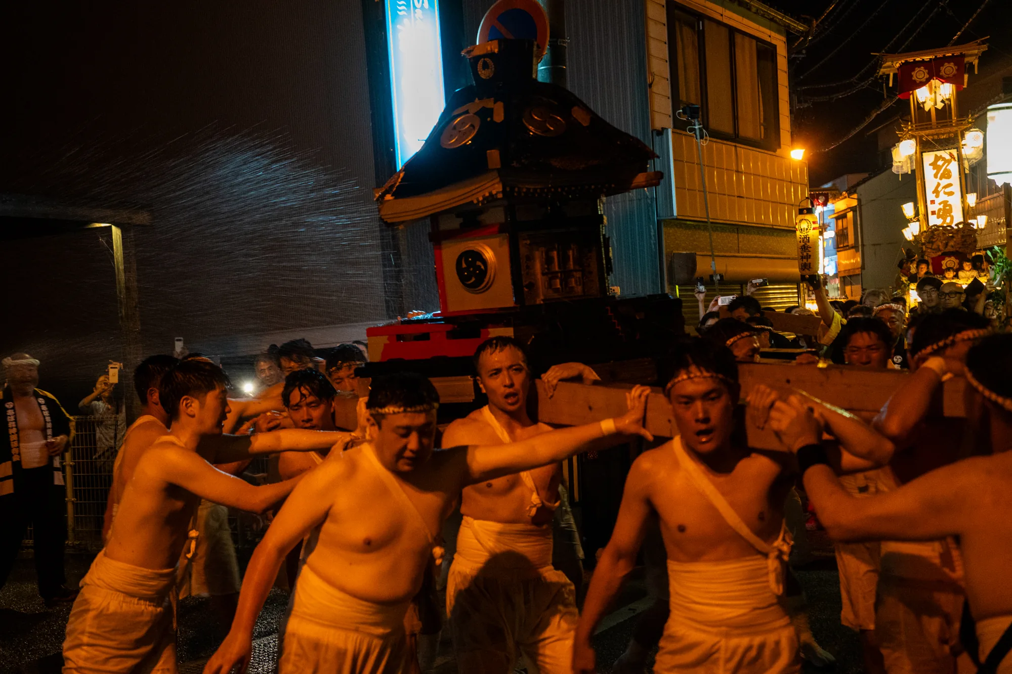 写真は日本の能登で撮影された祭りの様子を捉えています。夜の街を舞台に、ふんどし姿の若い男性たちが神輿を担いでいます。彼らの表情は真剣そのもので、額には汗が光っています。神輿は黒と朱色で装飾されており、重厚感があります。画面左側からは水が勢いよくかけられており、祭りの熱気をさらに高めています。背景には提灯や灯りが連なり、祭りの賑わいを伝えています。見物客も多く、スマートフォンで撮影している人も見られます。全体的に活気とエネルギーに満ち溢れた写真です。
