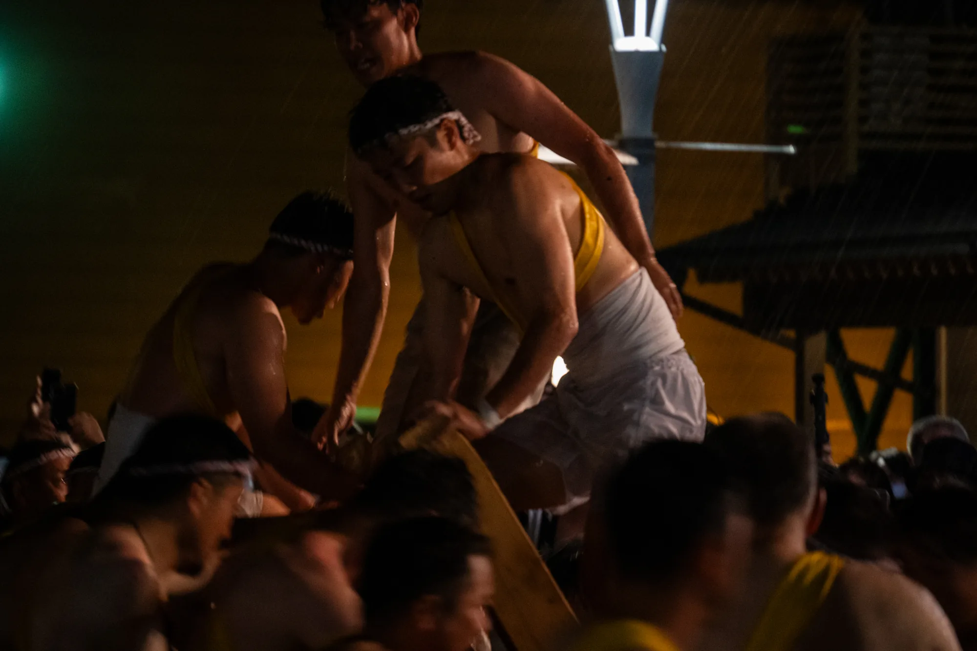 石川県能登で撮影された写真です。夜間の撮影で、雨が降っているようです。上半身裸で白い鉢巻を巻いた男性たちが複数写っており、祭りの様子を捉えています。黄色い肩掛けのようなものを身につけている人もいます。彼らは木製の何かを担いでいるようで、熱気と活気が感じられます。背景には街灯が見え、夜の闇を照らしています。全体的に暗いですが、雨に濡れた肌や力強い表情から祭りのエネルギーが伝わってくる写真です。