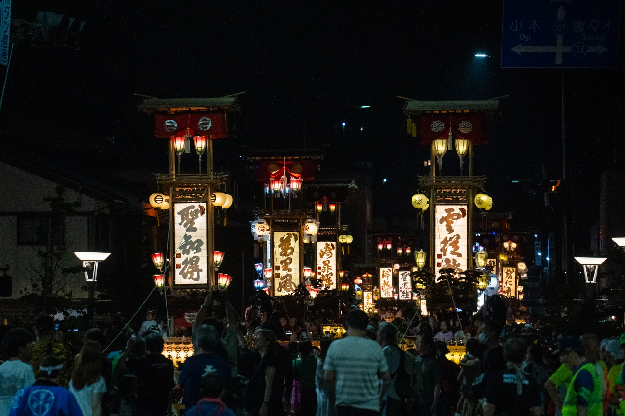 この写真は、日本の能登で撮影された夜のお祭り風景です。暗闇の中、多数の人々が集まっており、提灯や灯篭が明るく光を放っています。特に目を引くのは、漢字が書かれた大きな灯篭を載せた山車のようなもので、それぞれ異なる文字が書かれており、祭りの賑わいを象徴しています。これらの灯篭は、赤や黄色の提灯で飾られており、温かい光を周囲に投げかけています。群衆は、老若男女問わず、祭りの雰囲気を楽しんでいる様子が伺えます。背景には、道路標識が見え、「小木」「曽々木」といった地名が記されています。祭りの熱気と、伝統的な雰囲気が伝わってくる一枚です。