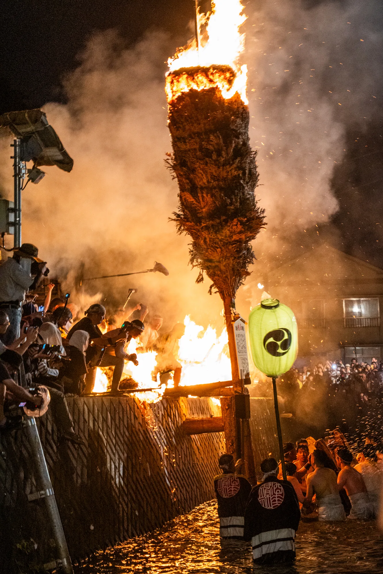 写真は、日本の能登地方で撮影されたと思われる、夜の火祭りの様子を捉えています。中央には、巨大な松明が燃え盛り、周囲には煙と火の粉が舞い上がっています。松明の根元には人々が集まり、祭りを盛り上げているようです。川の中に立ち、腰に白い布を巻いただけの男性たちが、松明に向かって何かをしている様子も見られます。堤防の上には多くの見物客が集まり、スマートフォンなどで写真を撮っています。画面右手には家屋が写り込み、祭りが行われている場所が地域社会の中心部であることがわかります。また、三つ巴の紋が入った提灯が立てられており、祭りの神聖な雰囲気を高めています。全体的に、力強く、熱気あふれる祭りの様子が伝わってくる写真です。