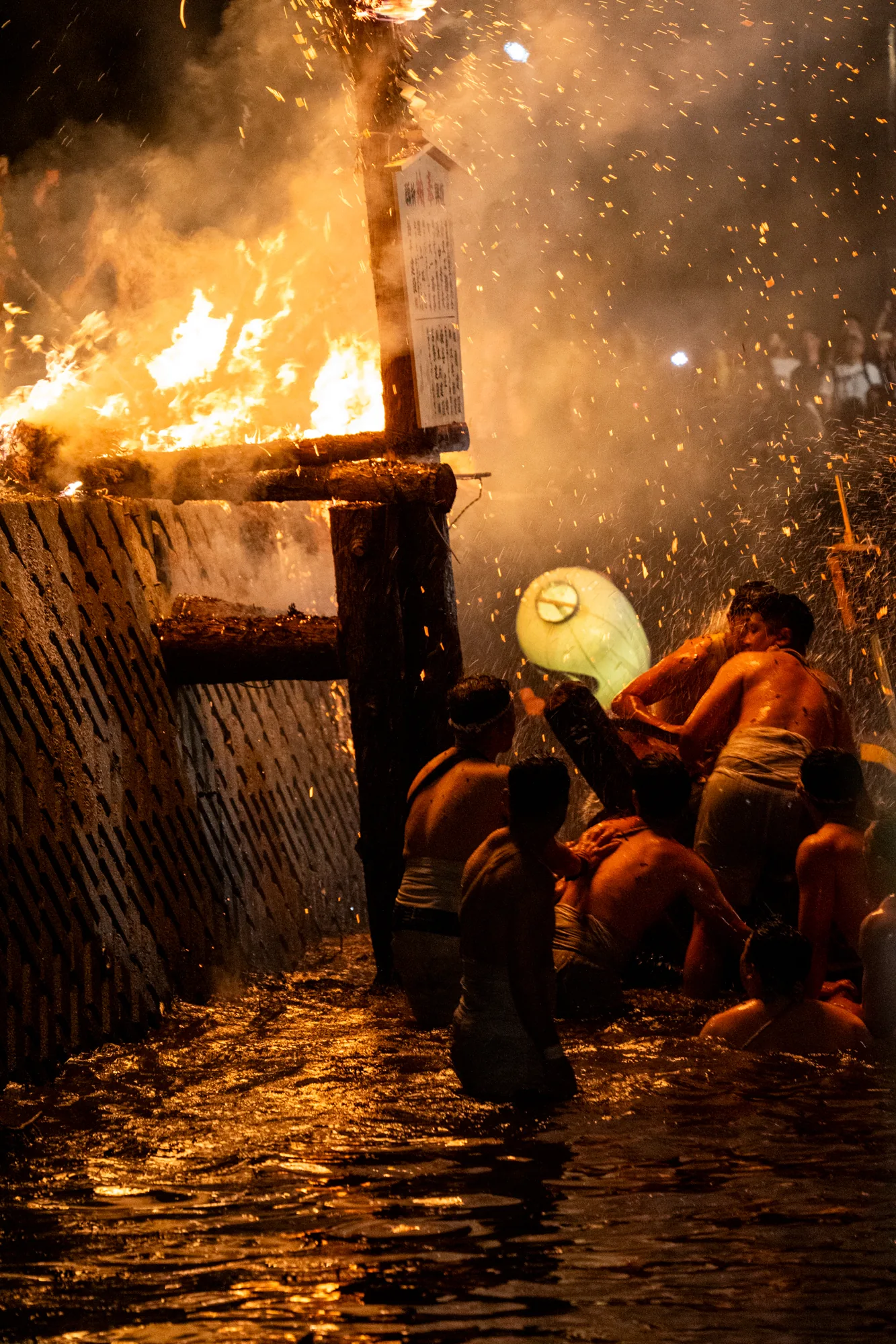 写真は日本の能登で撮影された火祭りの様子を捉えています。夜の暗闇の中、燃え盛る炎が強烈な光を放ち、周囲をオレンジ色に染め上げています。画面左側には木製の構造物が燃えており、その横には札のようなものが立てられています。炎からは大量の煙が立ち上り、火の粉が舞い散っています。  画面右側には、ふんどし姿の男性たちが水の中に立っており、何かの木材を担いでいます。水面は炎の光を反射して金色に輝き、水しぶきが勢いよく上がっています。彼らの表情は捉えられていませんが、その身のこなしから祭りに参加する熱意と力強さが伝わってきます。奥には群衆の姿も見え、祭りの活気が感じられます。  全体的に、熱気と興奮に満ちた、臨場感あふれる一枚です。炎の力強さ、水しぶきの躍動感、そして人々の熱気が混ざり合い、見る者に強い印象を与えます。能登の伝統的な祭りの様子を鮮やかに切り取った写真と言えるでしょう。