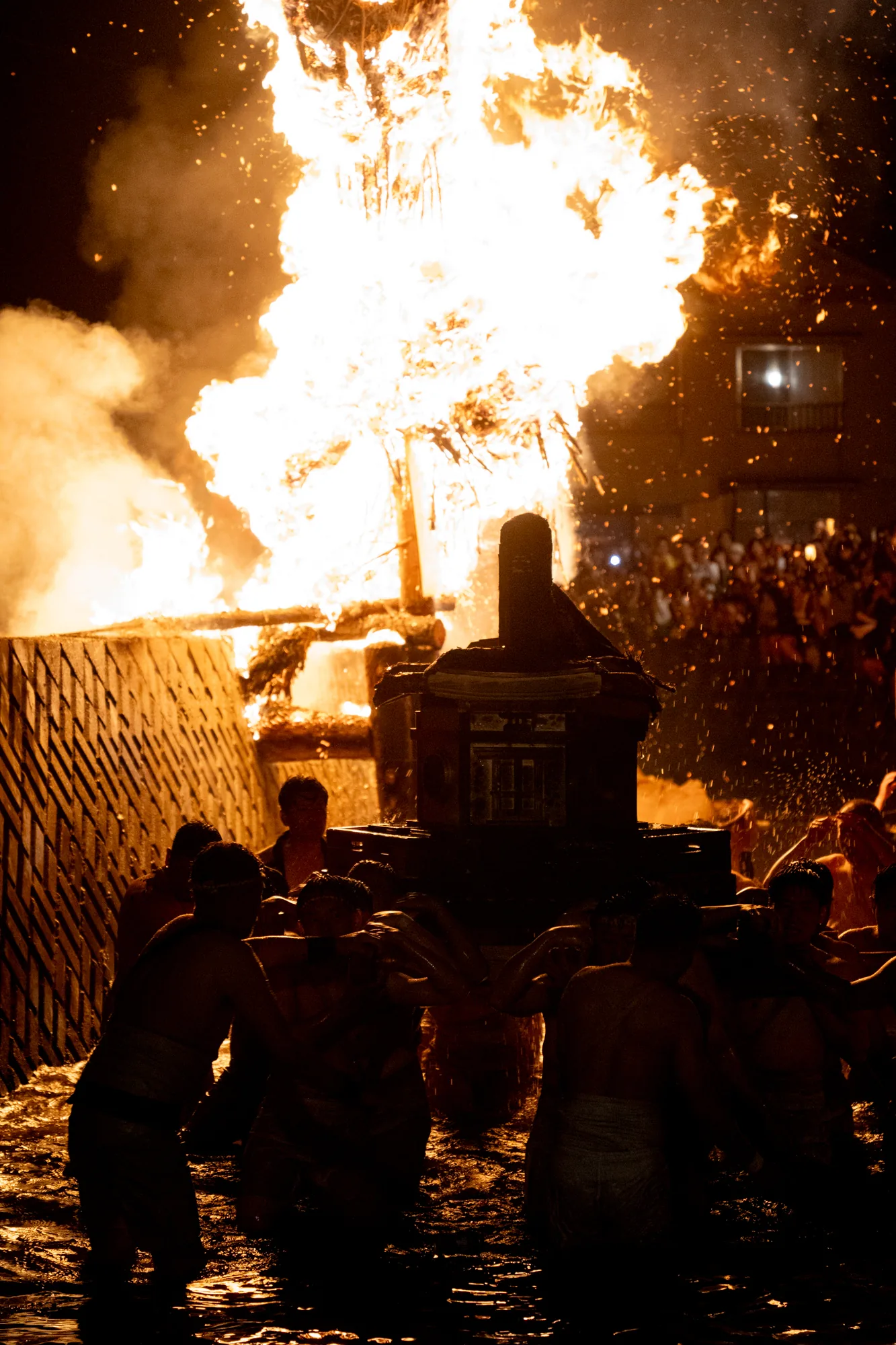 写真は、日本の能登で撮影されたと思われる火祭りの様子を捉えています。燃え盛る巨大な炎が画面上部を占め、その下では、多くの人々が担いでいると思われる神輿のようなものがシルエットで浮かび上がっています。担ぎ手たちは水中に浸かっており、水面が光を反射して輝いています。炎の熱気と水しぶきが混ざり合い、祭りの熱狂とエネルギーが伝わってくるようです。背景には群衆のシルエットも見え、窓の明かりがぼんやりと見えています。全体的に暗いトーンですが、炎のオレンジ色が強烈なコントラストを生み出し、見る者に強い印象を与えます。祭りの躍動感、力強さ、そして神聖さが凝縮された一枚と言えるでしょう。