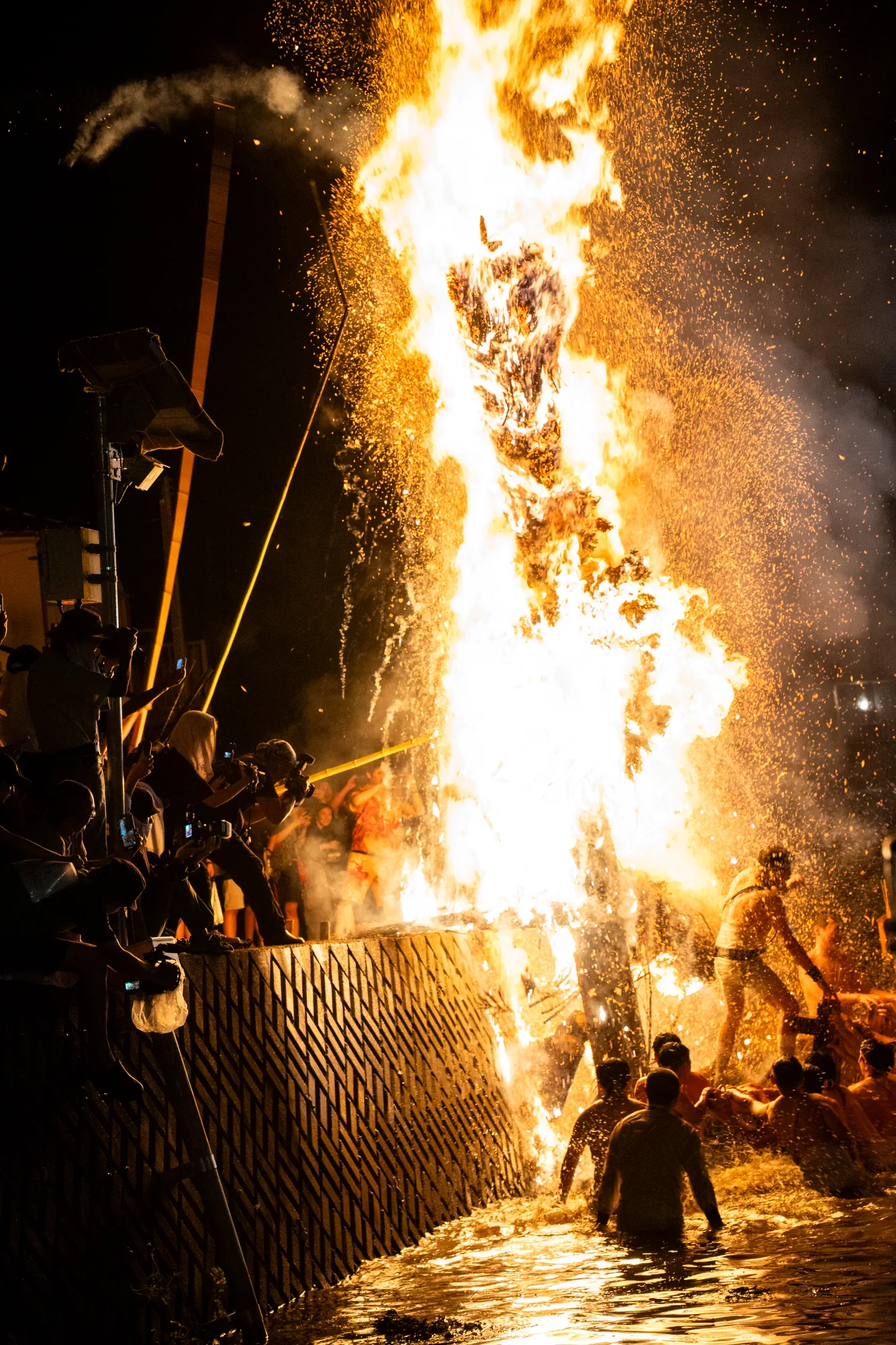 日本の能登で撮影された夜の写真です。中央には巨大な炎が燃え盛っており、火の粉が周囲に飛び散っています。炎の前に人々が集まり、水に入って火に立ち向かっているように見えます。彼らは上半身裸で、伝統的な装いをしている人もいます。炎を撮影している見物人も多く、スマートフォンやカメラを構えています。写真全体は暗い色調ですが、炎の明るさが際立ち、祭りの熱気と興奮を伝えています。水面に炎が反射し、幻想的な光景を作り出しています。