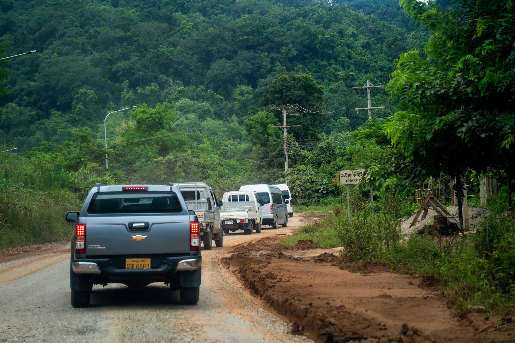 写真は、ラオスのルアンパバーンで撮影されたと思われる風景を捉えています。手前には、グレーのピックアップトラックが写っており、テールランプが点灯しています。ナンバープレートには「SI6661」と書かれています。トラックの前には、複数の小型トラックやバンが連なって走行しており、車列をなしています。路面は未舗装で、赤土がむき出しになっており、ぬかるんでいる箇所も見られます。道の脇には緑が生い茂り、熱帯の植生が豊かであることが伺えます。背景には、深い緑色の山がそびえ立ち、鬱蒼とした森林が広がっています。電柱と電線が森林の中に走っており、人々の生活の痕跡を感じさせます。道の脇にはラオス語と英語で書かれた「SHARP」という看板があり、現地の雰囲気を伝えています。全体的に、自然豊かなラオスの田舎道を、車がゆっくりと進んでいる様子が写し出されており、のどかで落ち着いた雰囲気が感じられます。