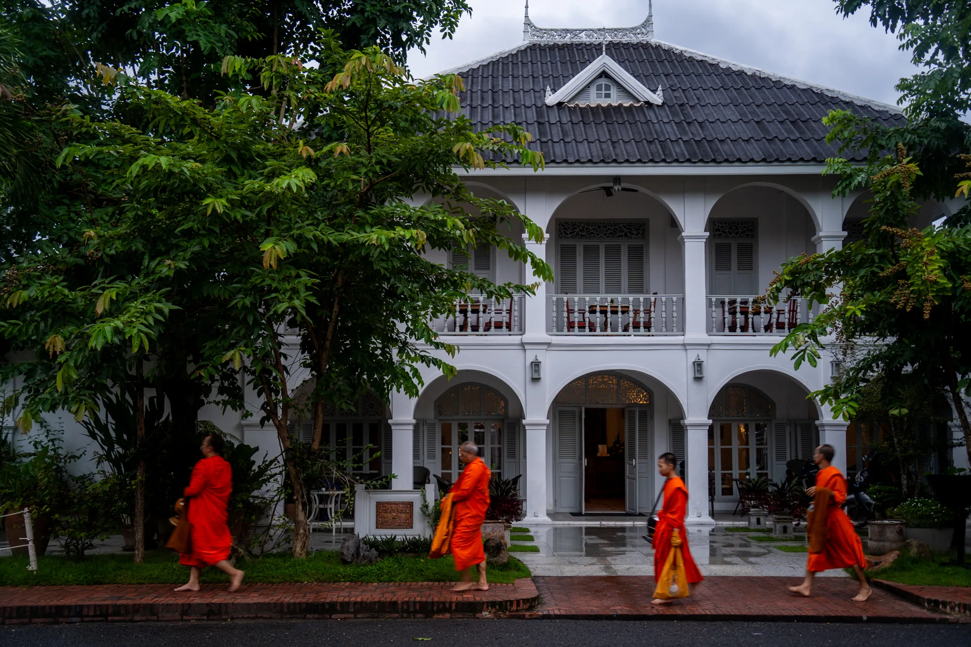 ラオスのルアンパバーンで撮影された写真です。画面中央には、白塗りの二階建てコロニアル様式の建物が写っています。建物はアーチ状の玄関とバルコニーを持ち、窓には白いルーバー式のシャッターが取り付けられています。屋根は黒い瓦葺きで、中央には小さな三角屋根の窓があります。建物の前には緑豊かな木々が茂り、建物を覆うように生い茂っています。  写真の左端から右端にかけて、鮮やかなオレンジ色の袈裟をまとった4人の僧侶が、舗装された道を裸足で歩いています。彼らは早朝の托鉢に向かうのでしょうか、あるいは寺院へ戻るのでしょうか。彼らの足元は、雨上がりで濡れているように見えます。空は曇りがちで、全体的に落ち着いた雰囲気の写真です。歴史的な建物と僧侶たちの姿が、ルアンパバーン独特の文化的な景観を表現しています。