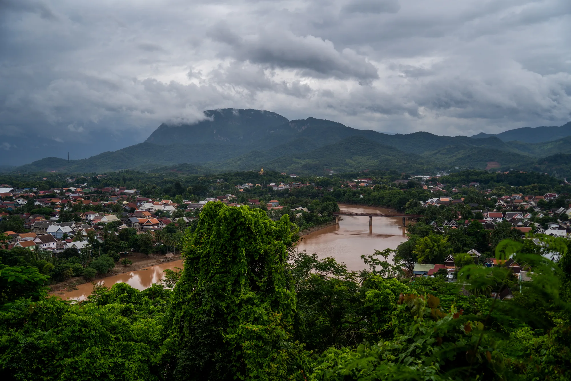 写真はラオスのルアンパバーンで撮影された風景です。画面全体は、緑豊かな熱帯の植生によって縁取られており、手前には濃い緑色の木々が茂っています。中央には、茶色く濁った川が蛇行しており、その上に橋が架かっています。川の両岸には建物が密集しており、屋根の色は赤茶色が目立ちます。家々の間には木々が点在し、緑と赤茶色のコントラストが美しい街並みを形成しています。遠景には、緑色の山々が連なり、山頂は雲に覆われています。空は厚い雲に覆われており、全体的にどんよりとした曇り空です。写真はややローアングルから撮影されており、奥行きのある構図になっています。全体的に落ち着いた雰囲気で、ルアンパバーンの自然と街並みが調和している様子が伝わってきます。