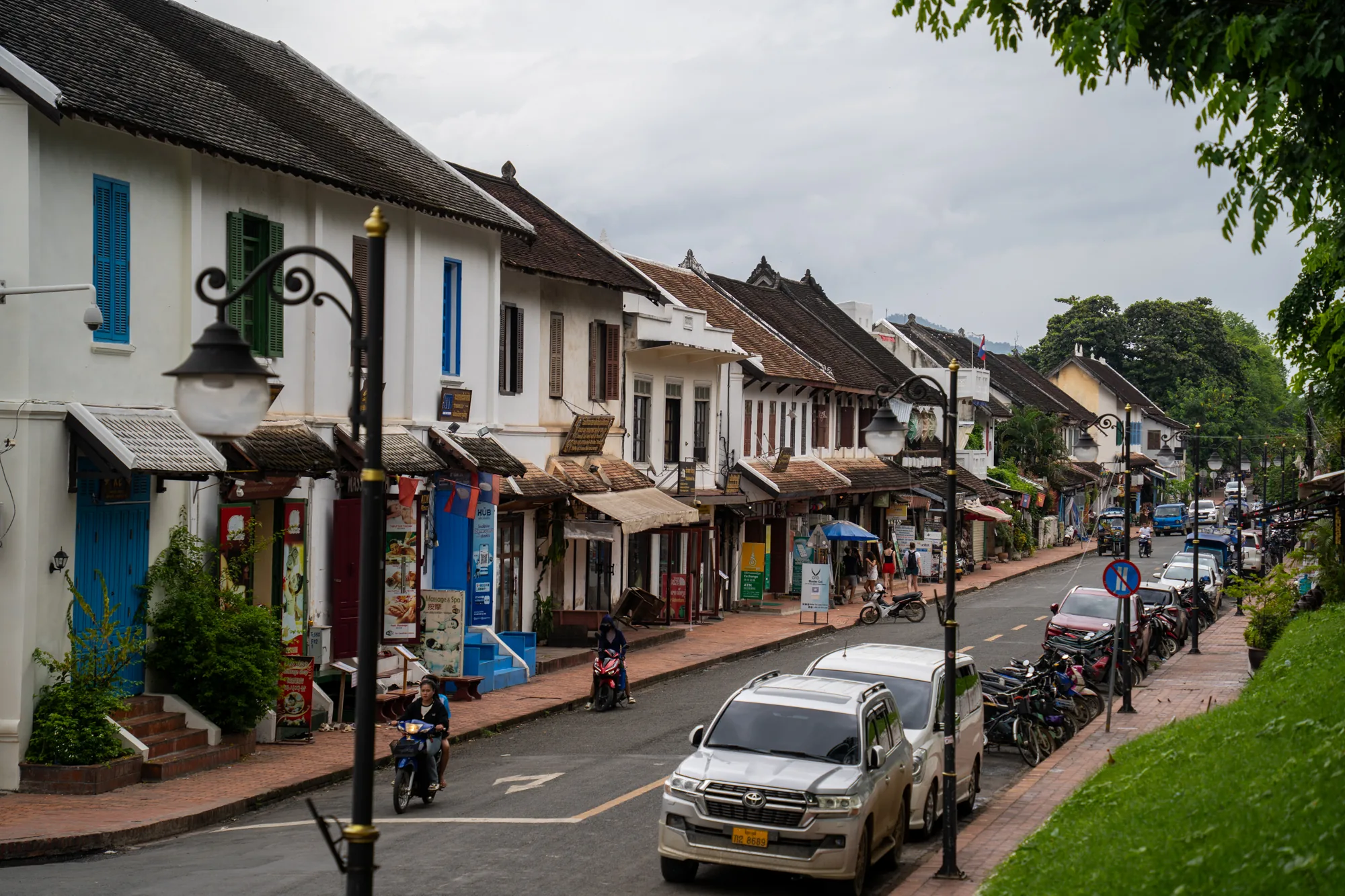 ラオスのルアンパバーンで撮影された写真です。画面奥へと続く道の両側に、コロニアル様式の建物が立ち並んでいます。これらの建物は白壁に茶色い屋根が特徴で、青や緑などカラフルな窓枠や扉がアクセントになっています。建物は店舗として使われているようで、看板や商品が見えます。道路には車やバイクが駐車されており、歩行者も数人見えます。手前にはレトロな街灯が立っており、道路の右側には緑の芝生があります。全体的に落ち着いた雰囲気で、ルアンパバーンの独特な街並みが表現されています。空は曇っており、穏やかな気候であることが伺えます。